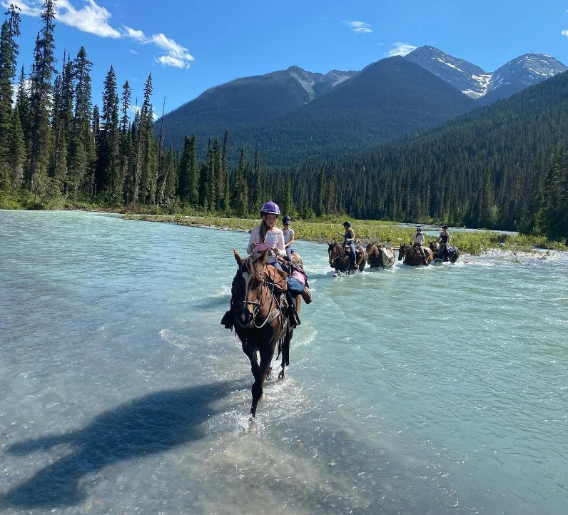 Horseback riders cross a turquoise river beneath forested, snow-capped mountains.
