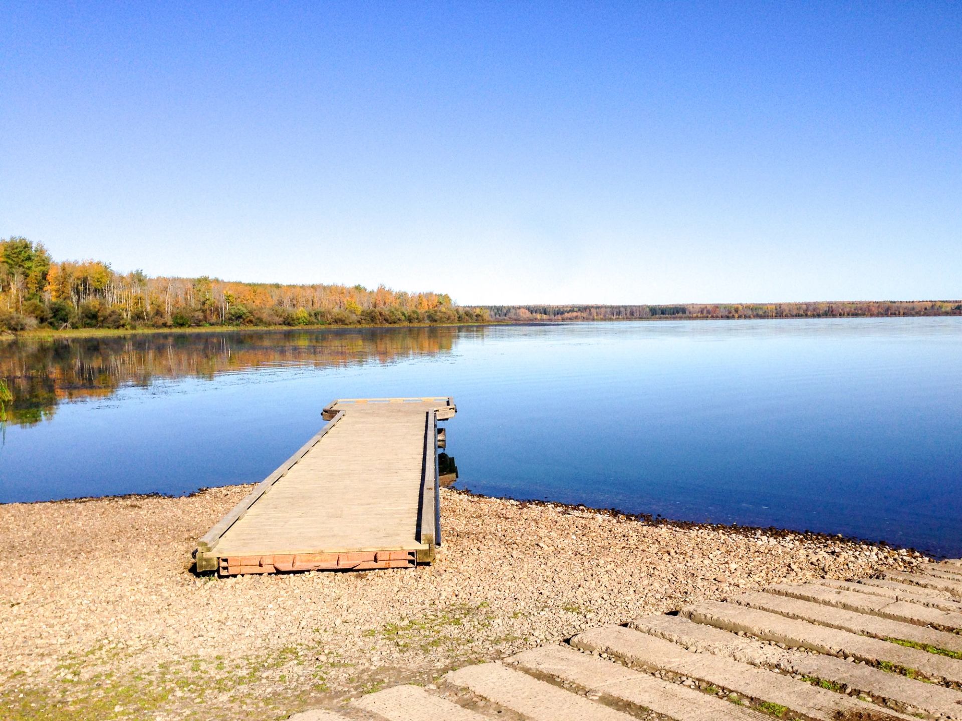 Wooden dock on clear lake with autumn trees and blue sky.