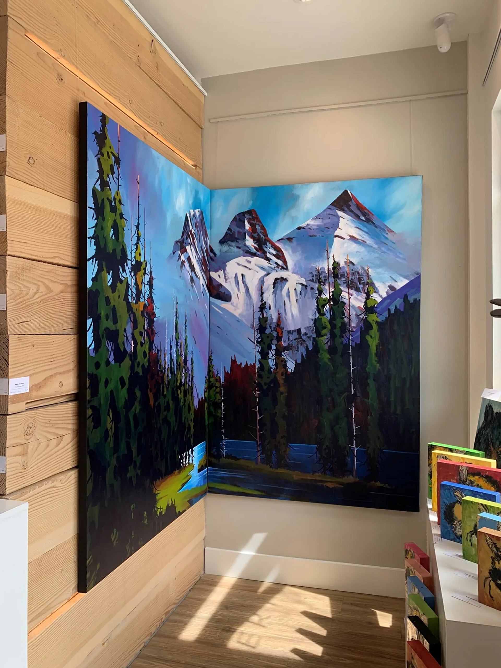 A gallery corner displays two vibrant paintings that merge to create a panoramic mountain and forest scene.