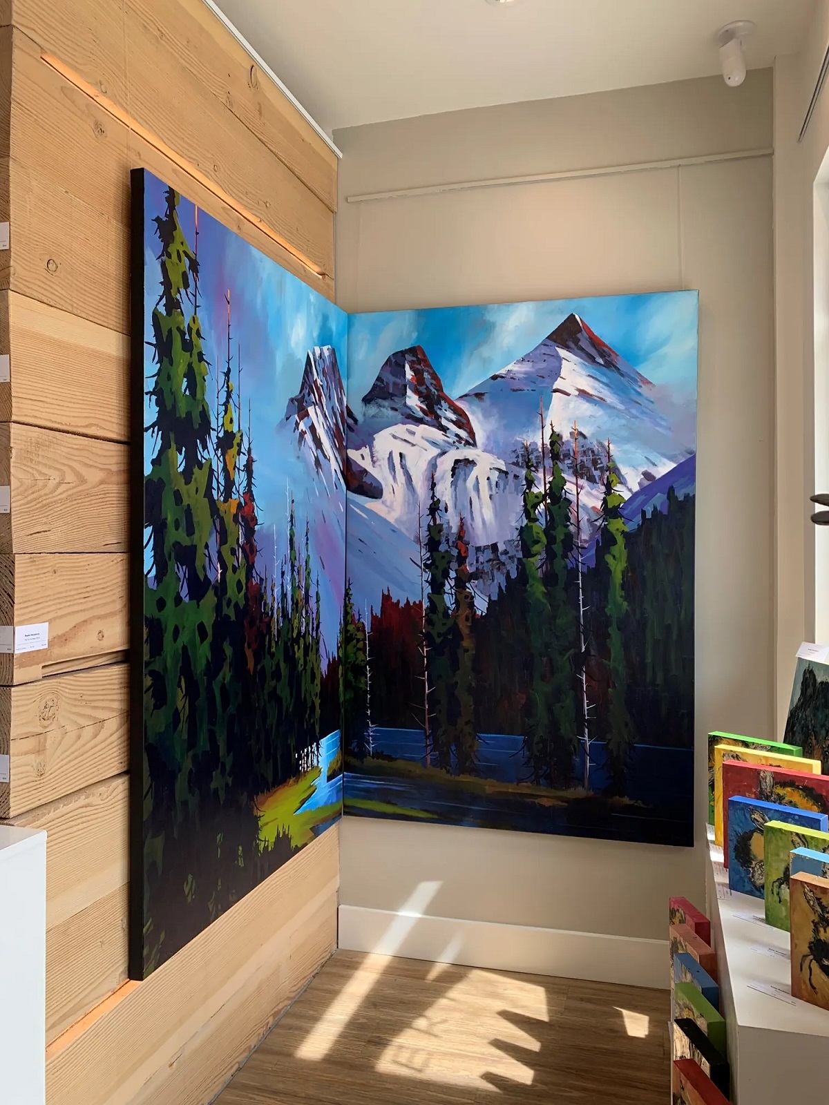 A gallery corner displays two vibrant paintings that merge to create a panoramic mountain and forest scene.