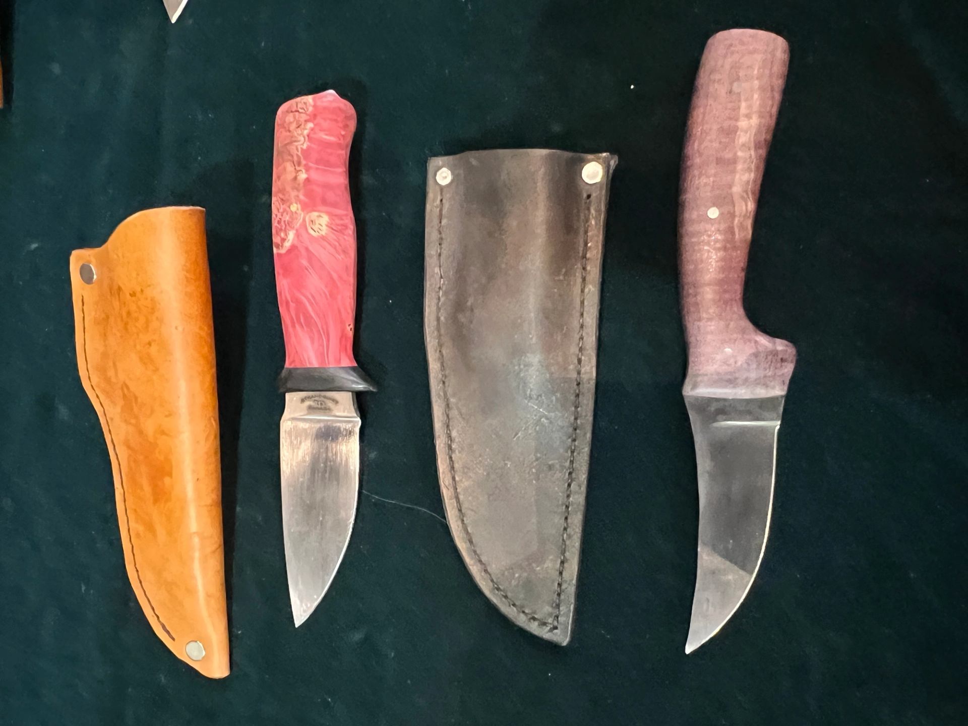 Knives with coloured handles and leather sheaths laid on black fabric.