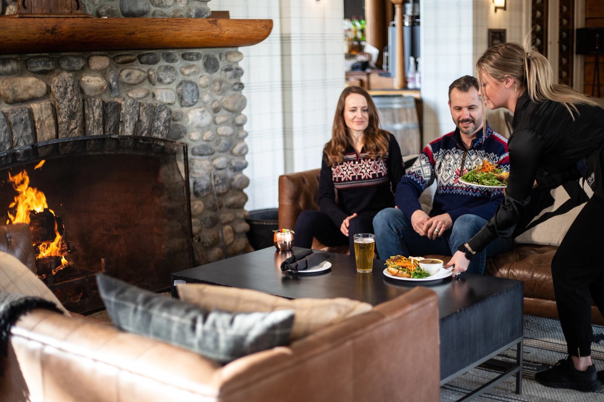 Two people seated on leather chairs as a server places food near a glowing stone fireplace