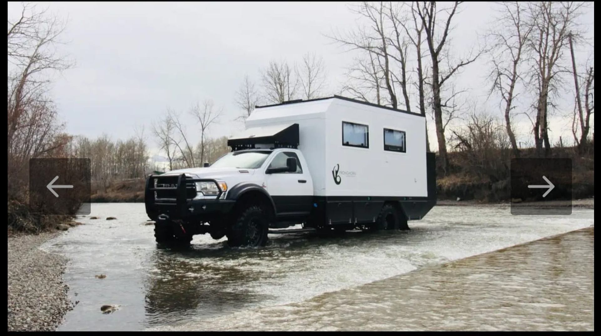Off-road expedition truck crossing shallow river in rugged, leafless terrain.