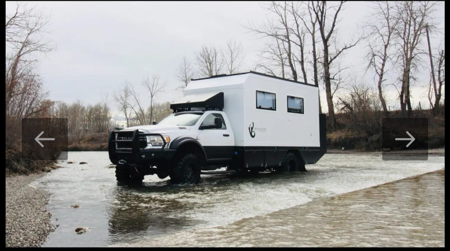 Off-road expedition truck crossing shallow river in rugged, leafless terrain.