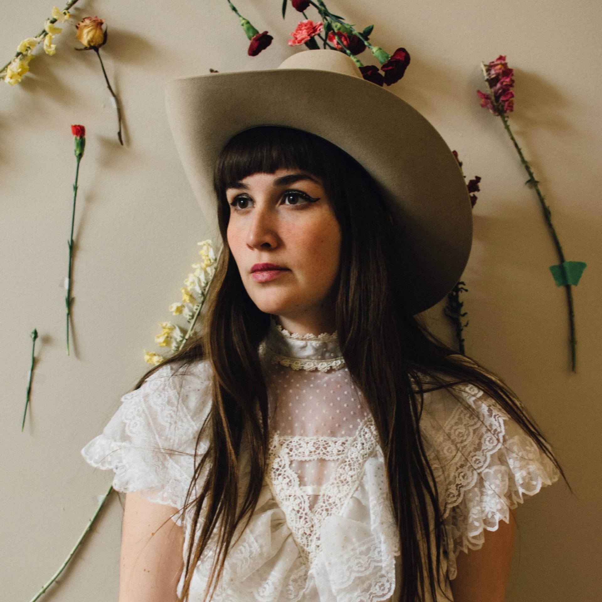 Person in a cowboy hat and lace top standing against a wall decorated with flowers.