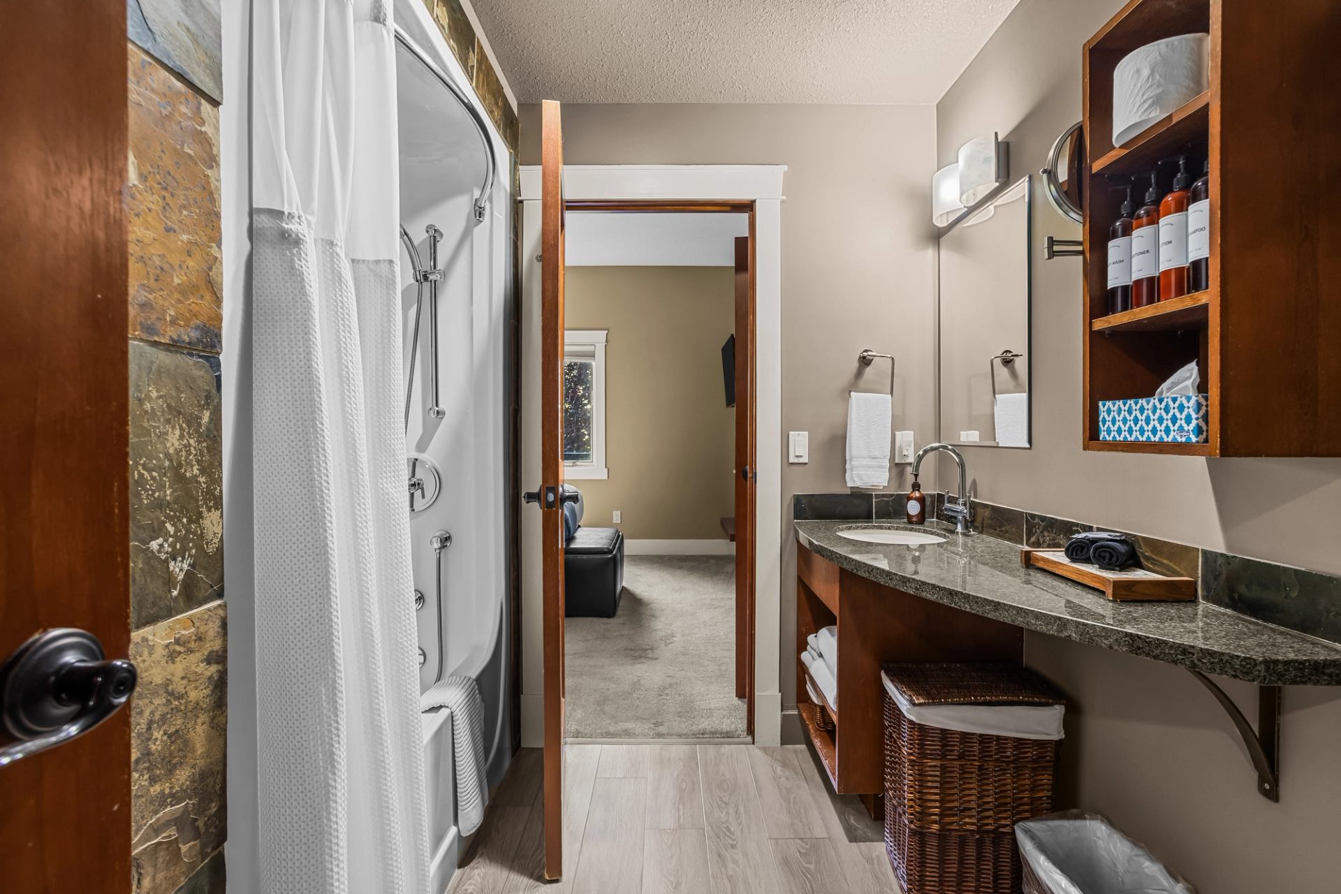 Bathroom with shower, granite vanity, mirror and wooden shelves stocked with toiletries.