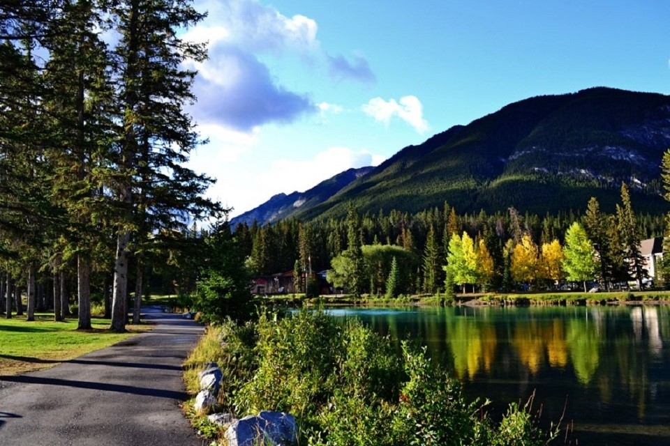 Paved trail beside calm river, trees and mountains reflected in water.