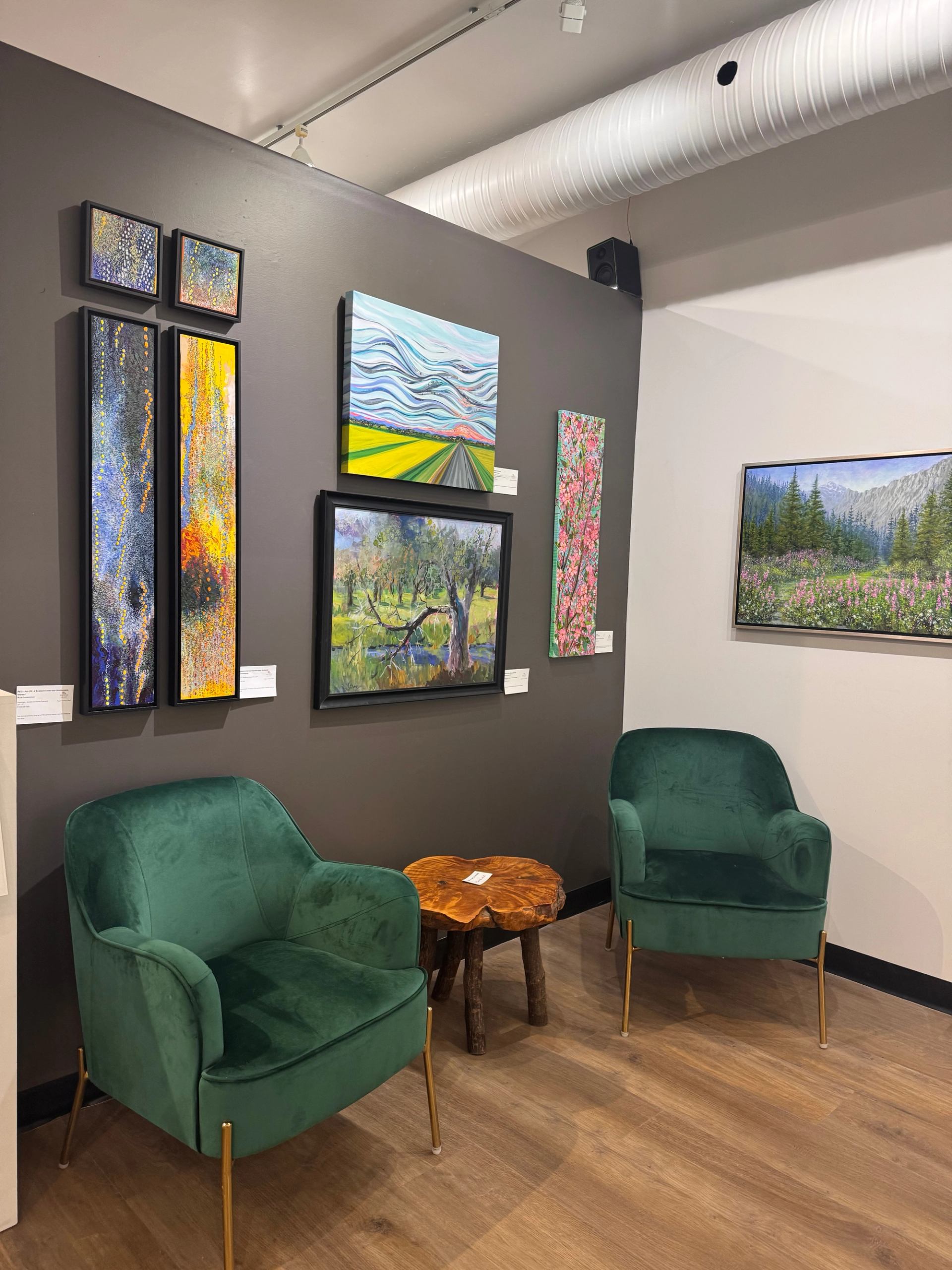 Green armchairs and a wood table sit beneath colourful paintings in an art gallery.