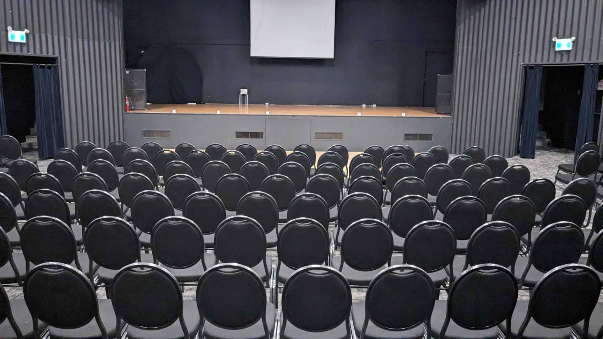 Rows of empty black chairs facing a small stage with screen and speakers.