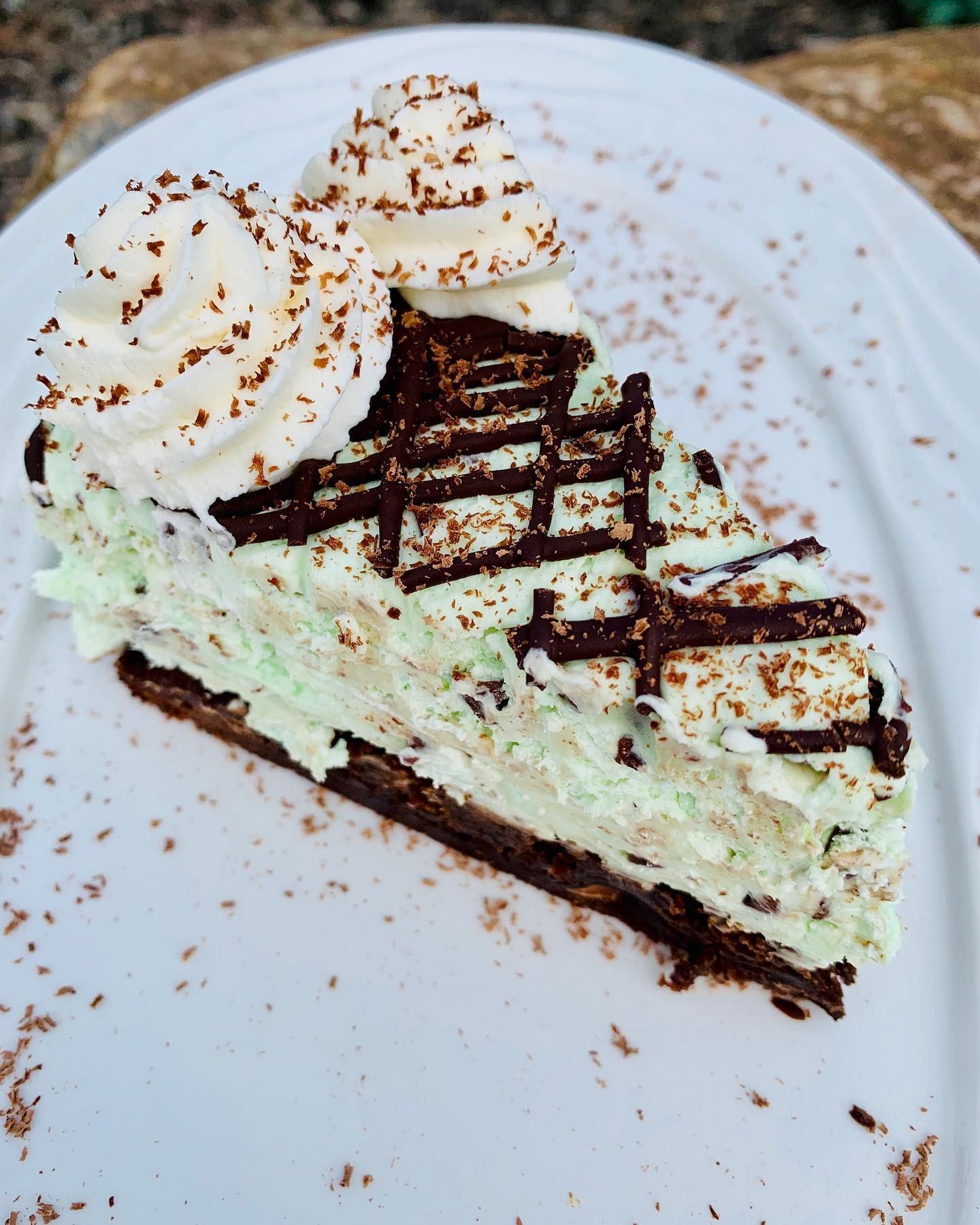 Slice of layered green dessert with whipped cream, chocolate drizzle and shavings on plate.