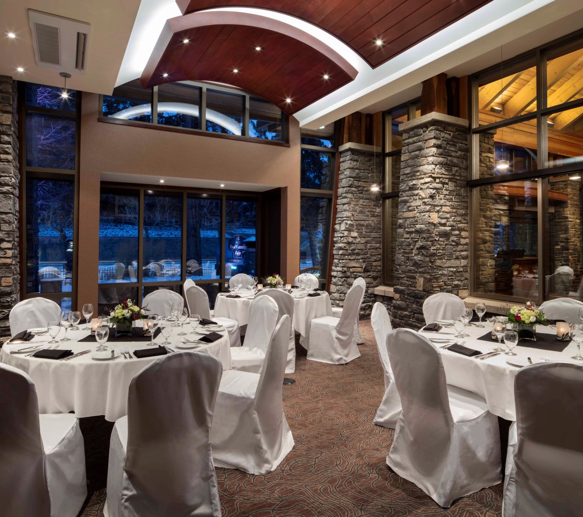 Banquet room with round tables, white chair covers, stone pillars and tall windows.