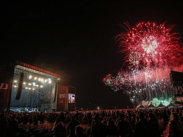 Crowd at outdoor concert watches large red and white fireworks burst over main stage.