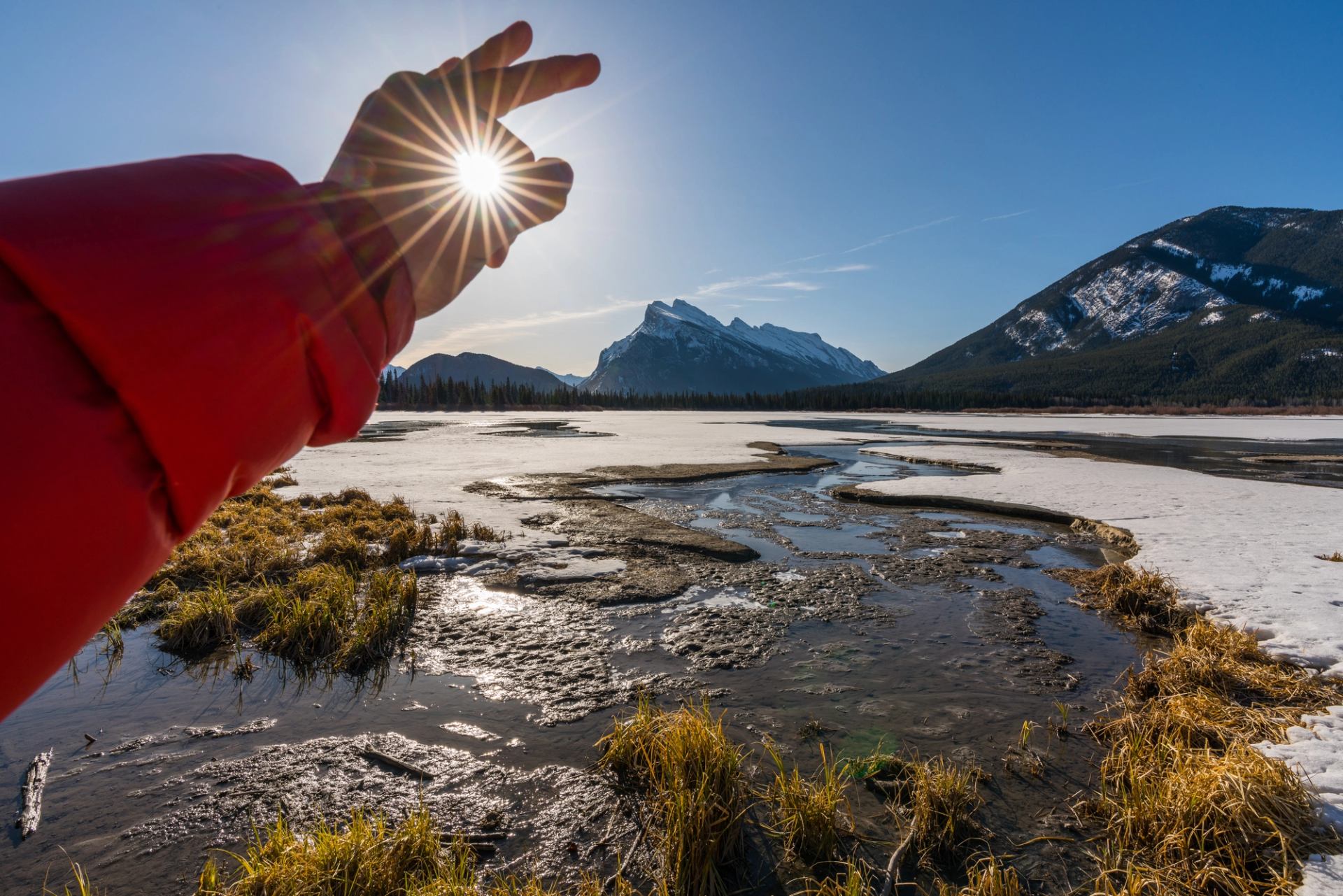 Hand framing the sun above frozen wetlands with snow-capped peaks in the distance.