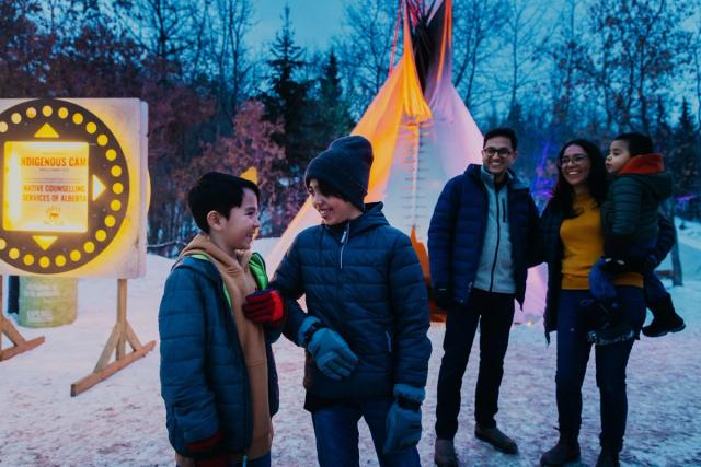 Two boys smile while standing in front of a tipi on a snowy day.