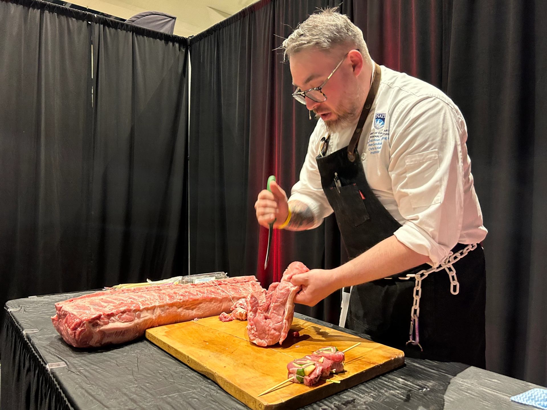 Chef in apron trims large cut of raw meat on wooden cutting board.