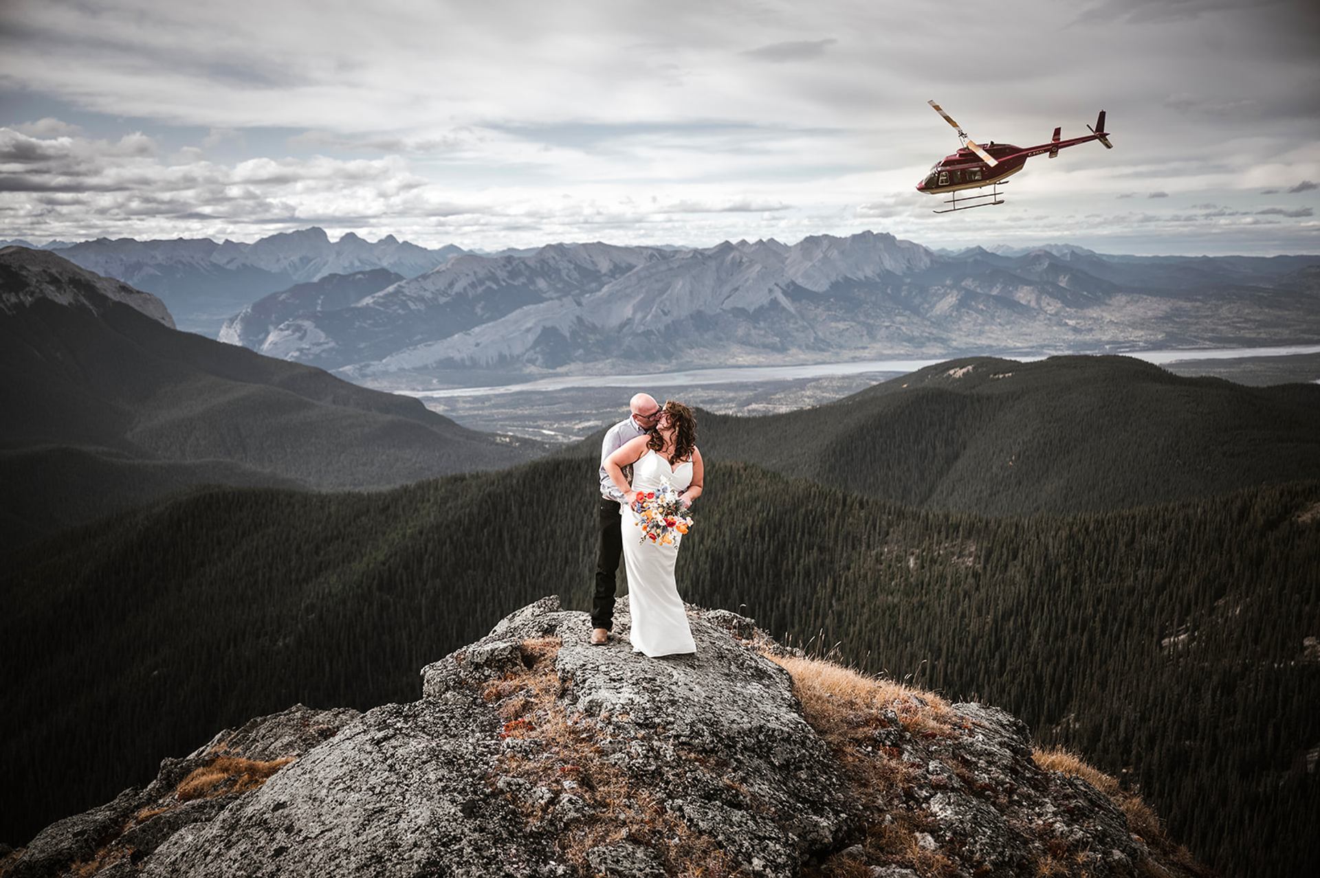 A couple embraces on a rocky peak while a helicopter hovers nearby.