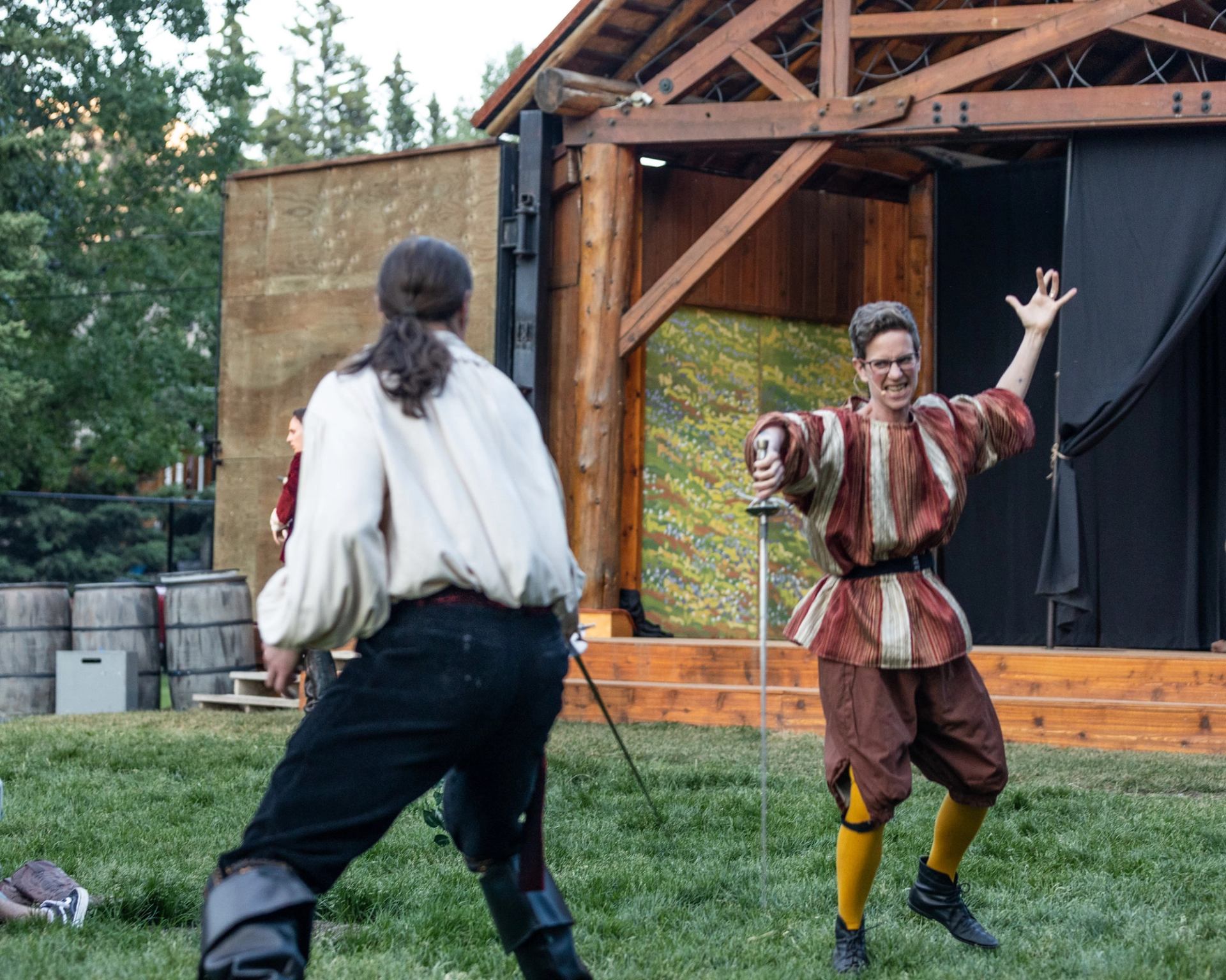 Two actors in period costumes perform a sword fight on an outdoor stage during a live theatre show at the Stan Rogers Stage in Canmore.