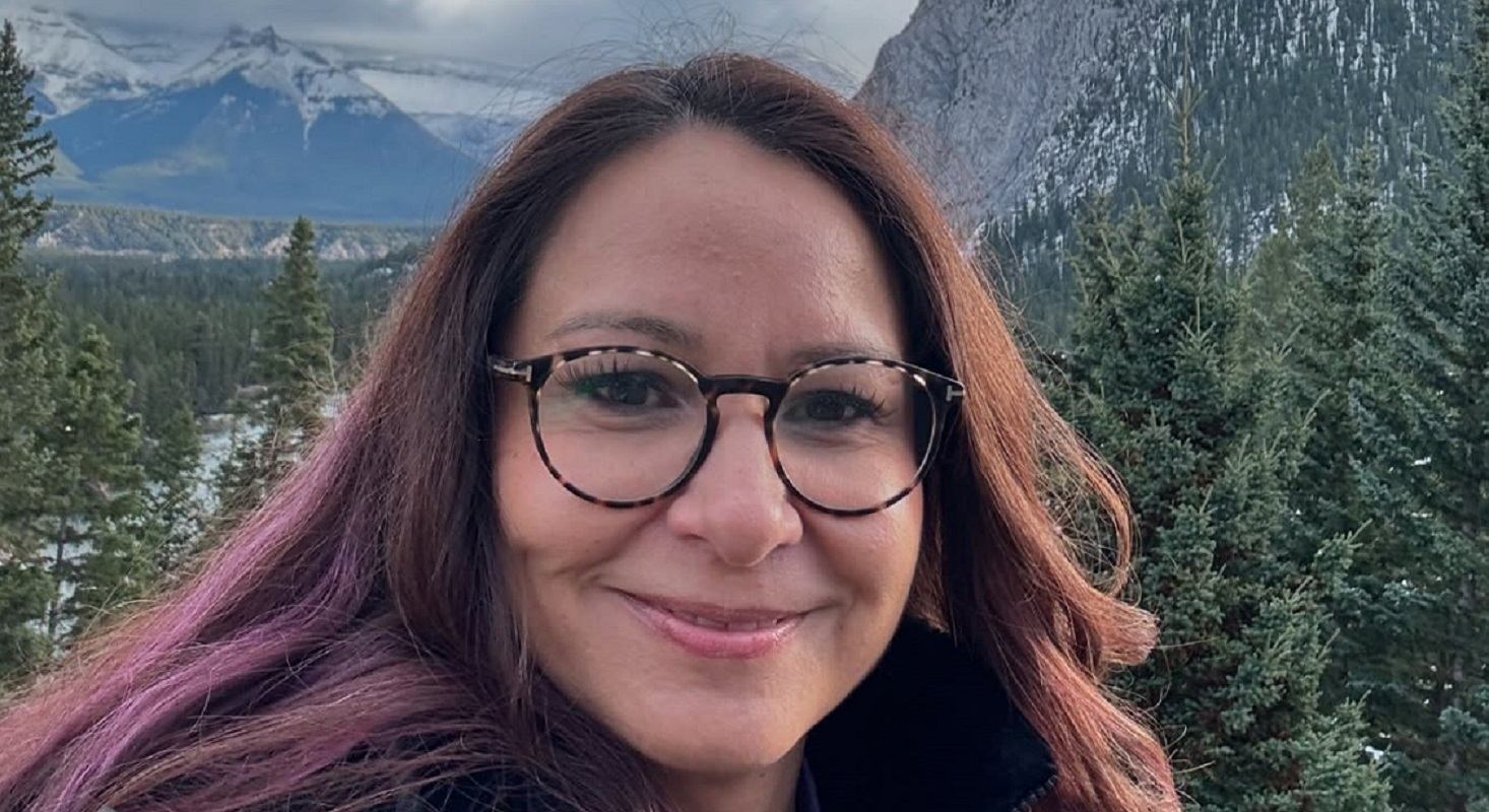 Person wearing glasses stands before evergreen trees and snow-capped mountains in the distance