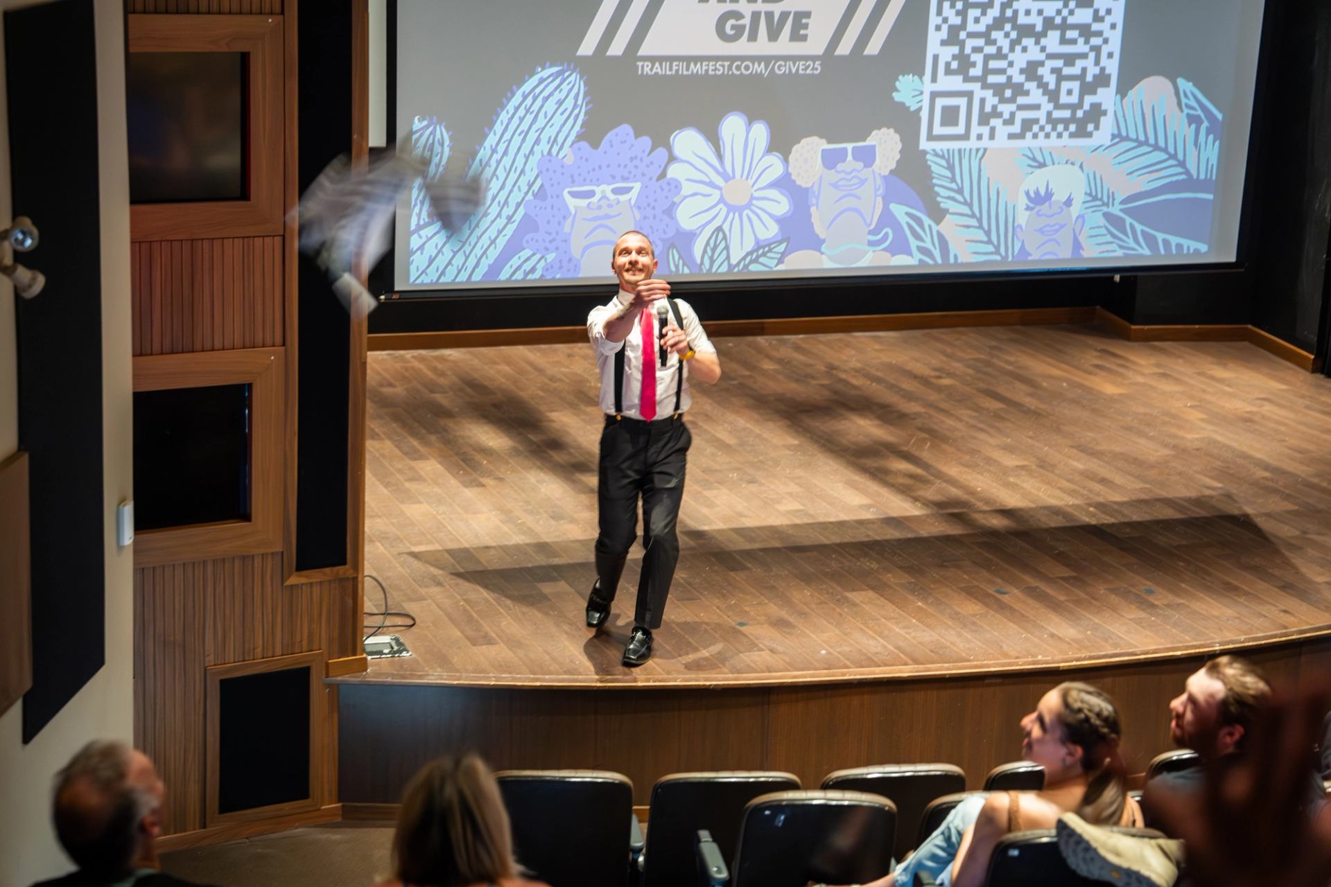 Presenter on theatre stage throws object toward seated audience before large projection screen.