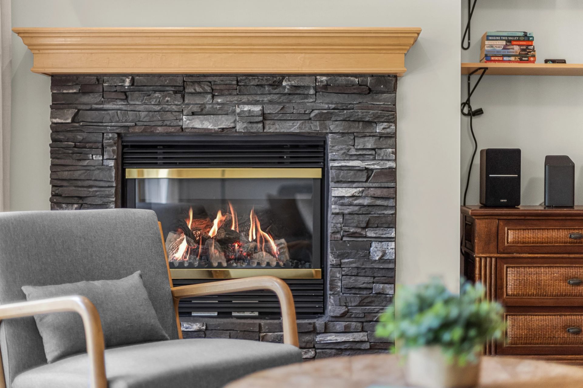 Stone fireplace with burning logs beside a grey armchair and wooden shelving.