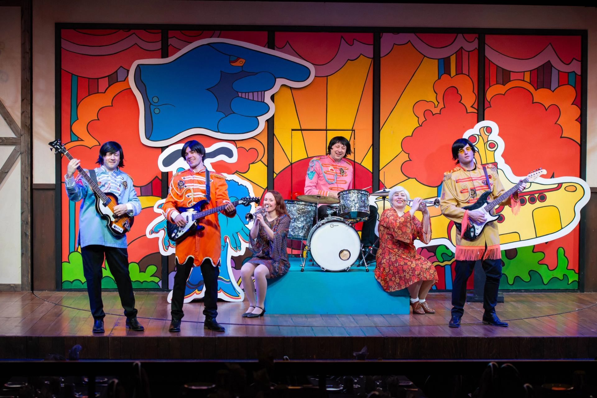 Stage performers in colourful Sgt. Pepper costumes play instruments before psychedelic cartoon backdrop.
