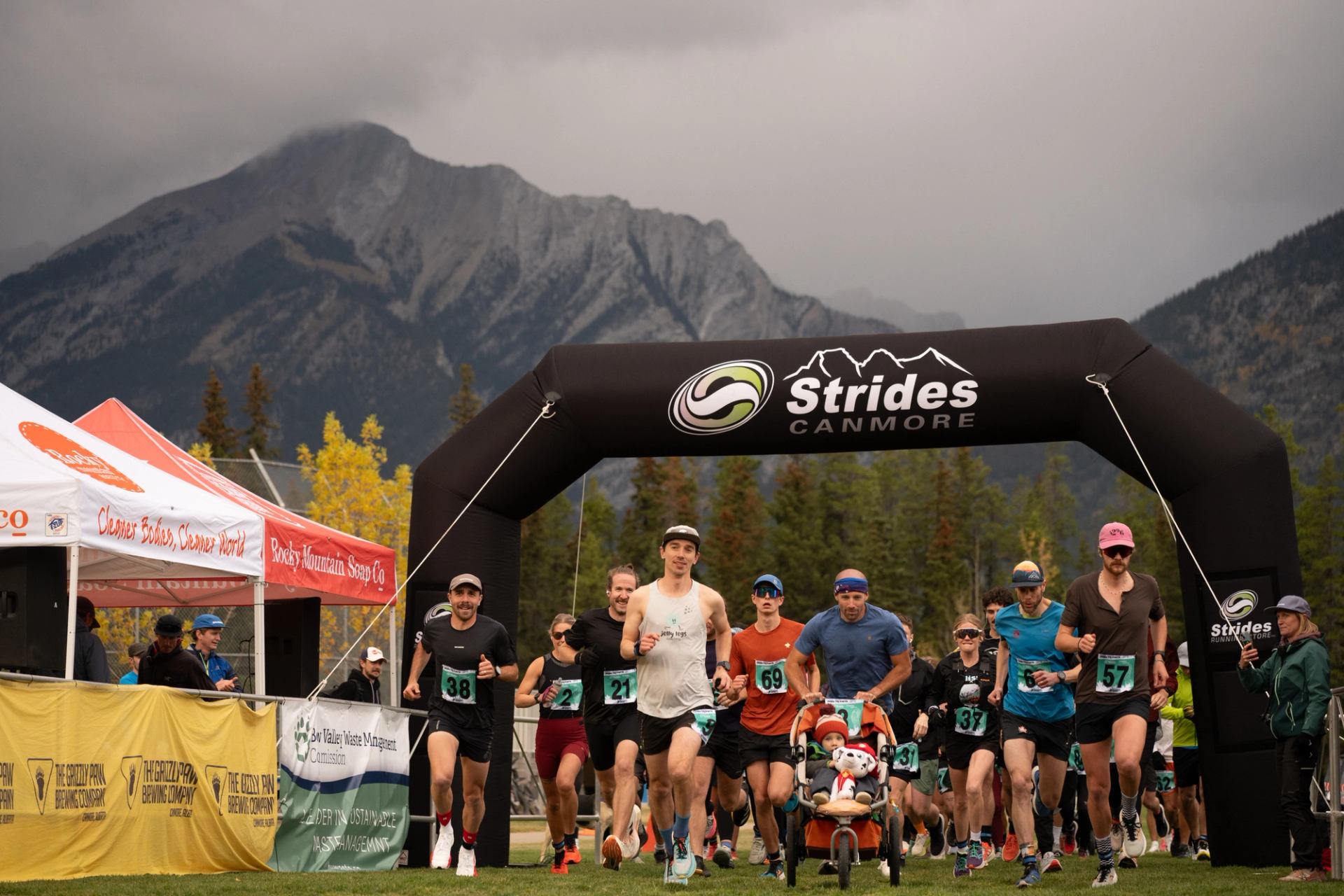Runners start a race under Strides Canmore arch with mountains and trees behind.