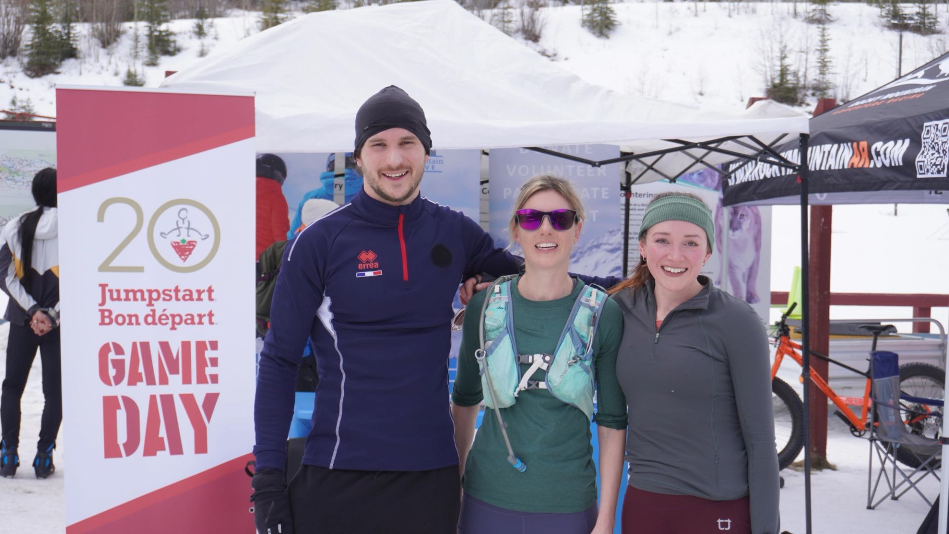 Three people posing together at a winter event near a Game Day sign and tents.