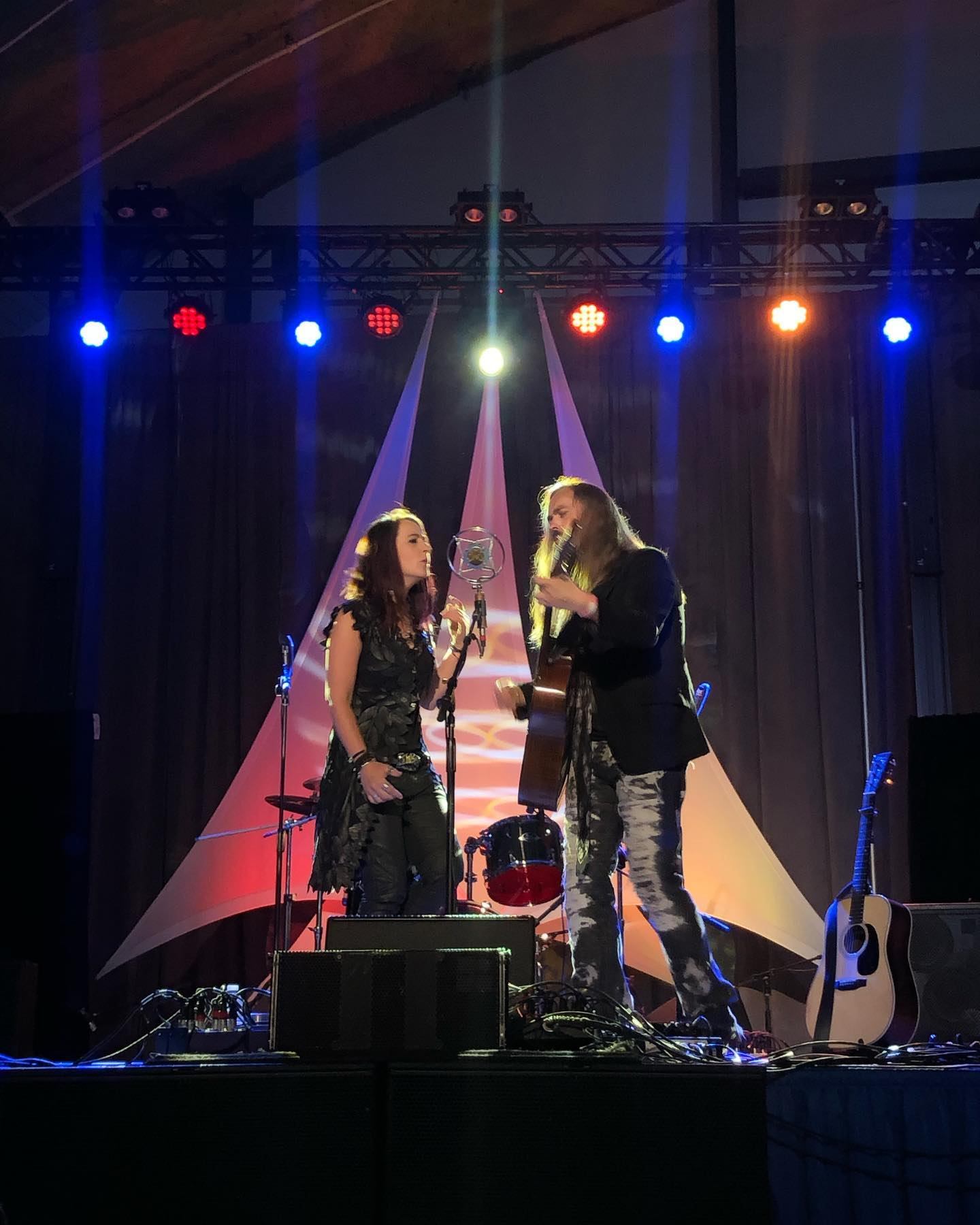 Two musicians sing into a vintage microphone on stage with guitars and coloured lights.