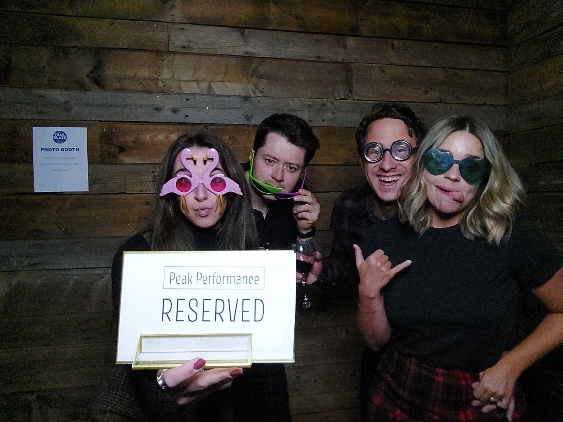 Eight people in novelty sunglasses pose with playful signs in front of a wooden wall.