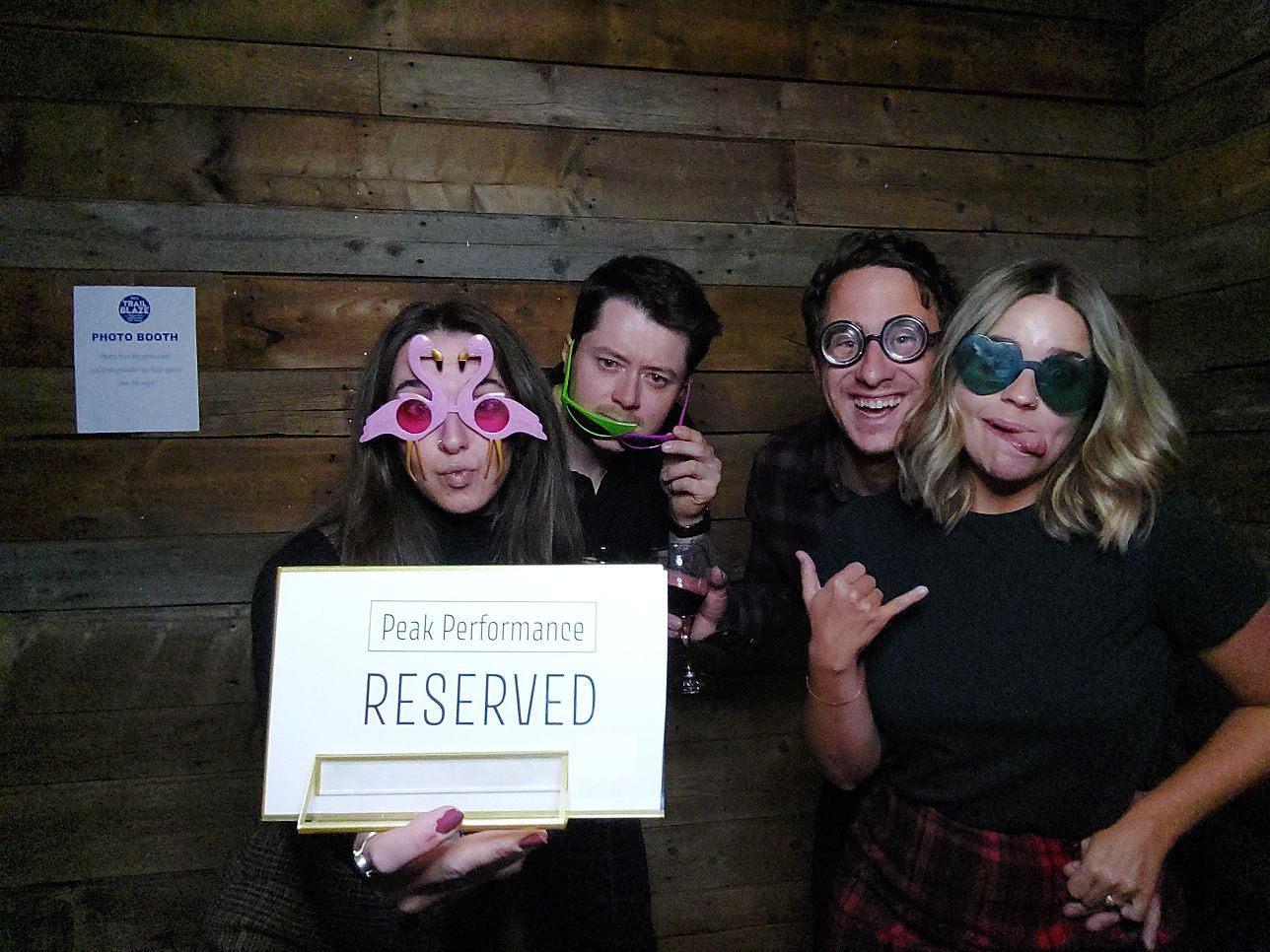 Eight people in novelty sunglasses pose with playful signs in front of a wooden wall.