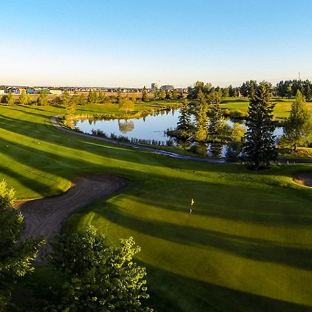 Golf course with pond, bunkers, and trees under soft morning or evening light.