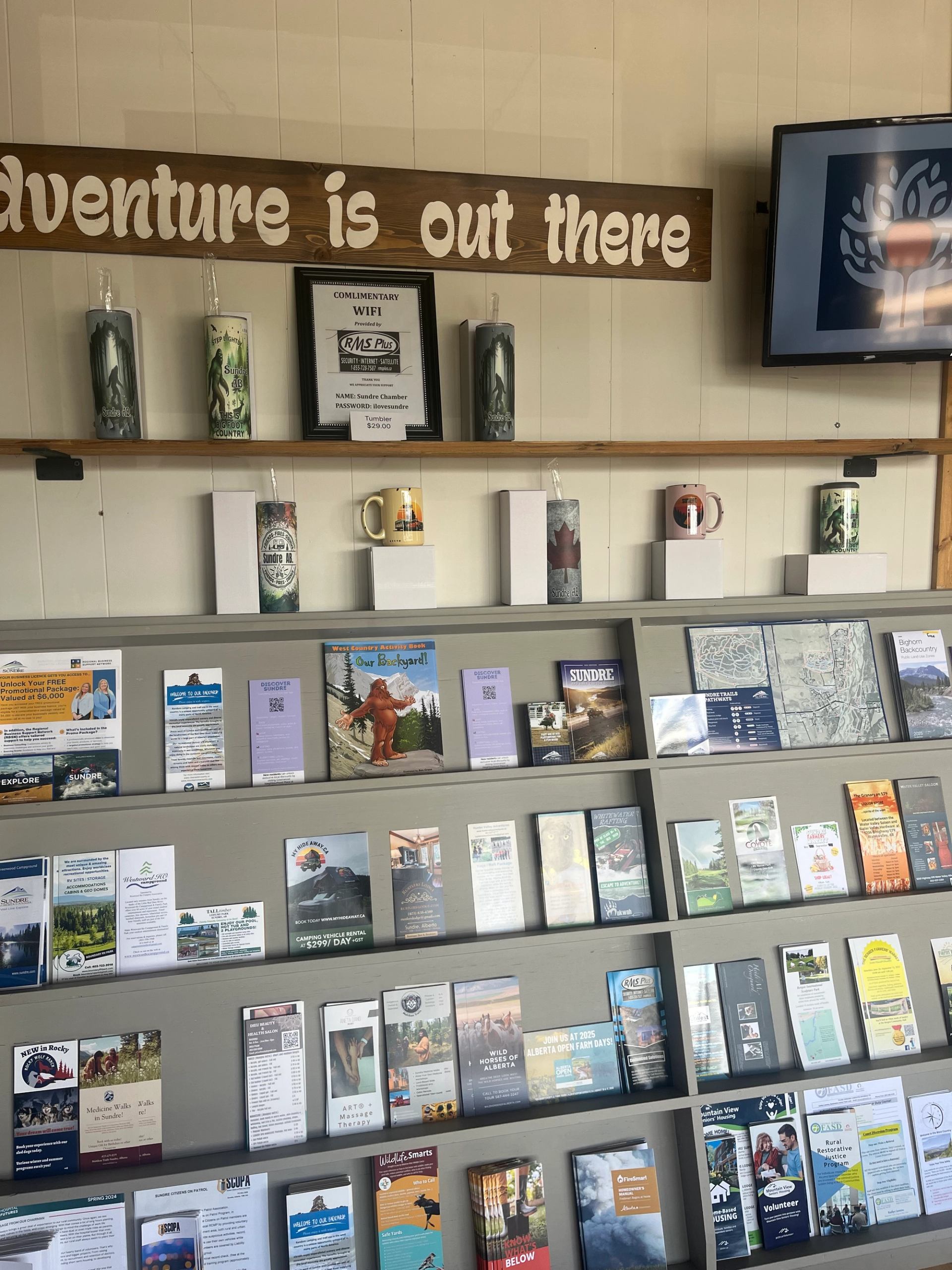 Sundre Visitor Centre wall with brochures, containers, and a TV under “Adventure is out there” sign.