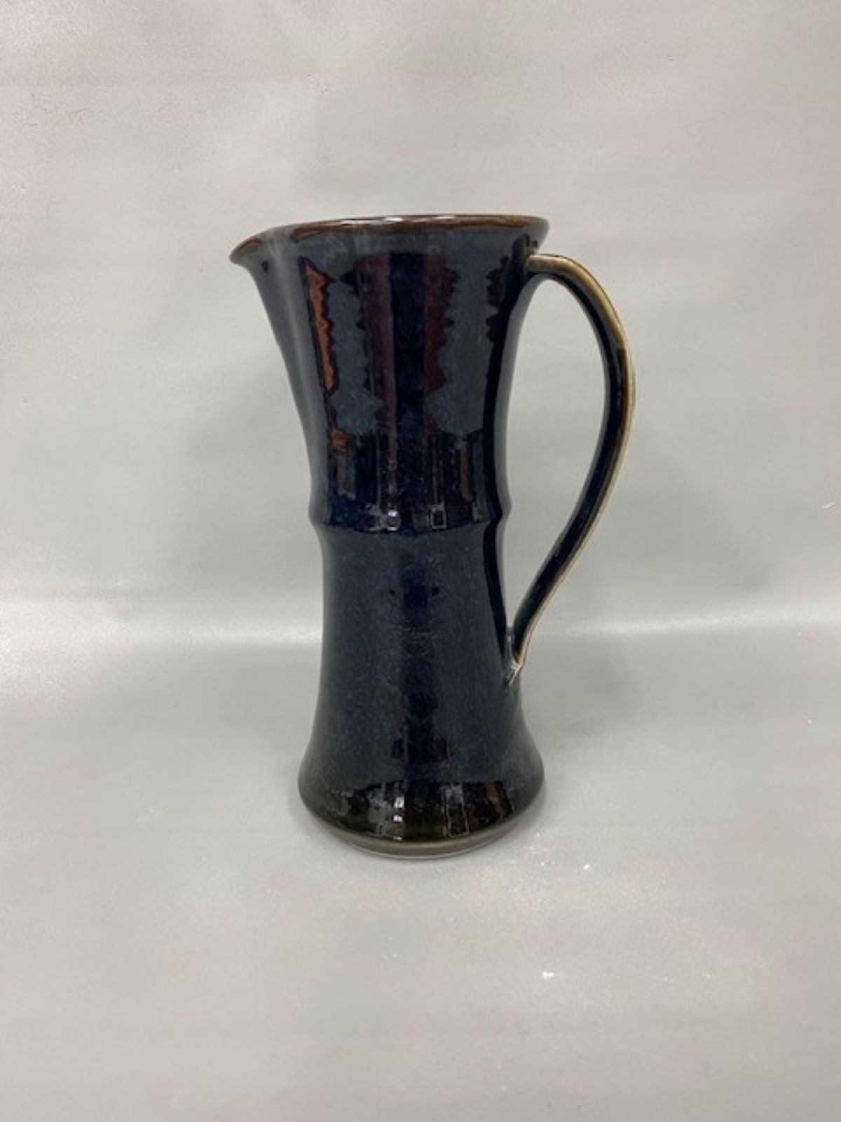 Tall handmade ceramic pitcher with a curved handle and dark blue glaze on a neutral background.