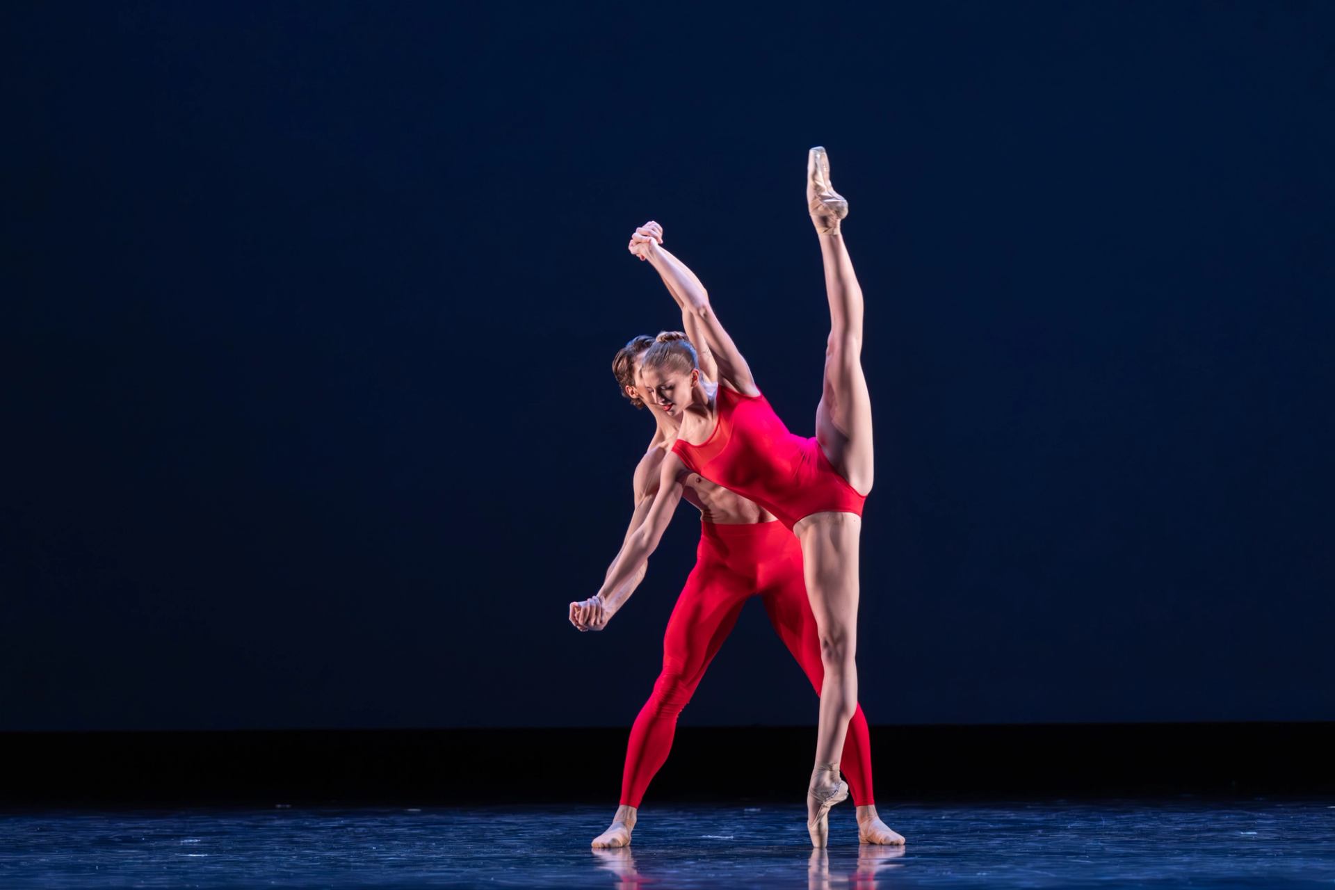Two ballet dancers dressed in red. The ballerina has her leg posed straight up.