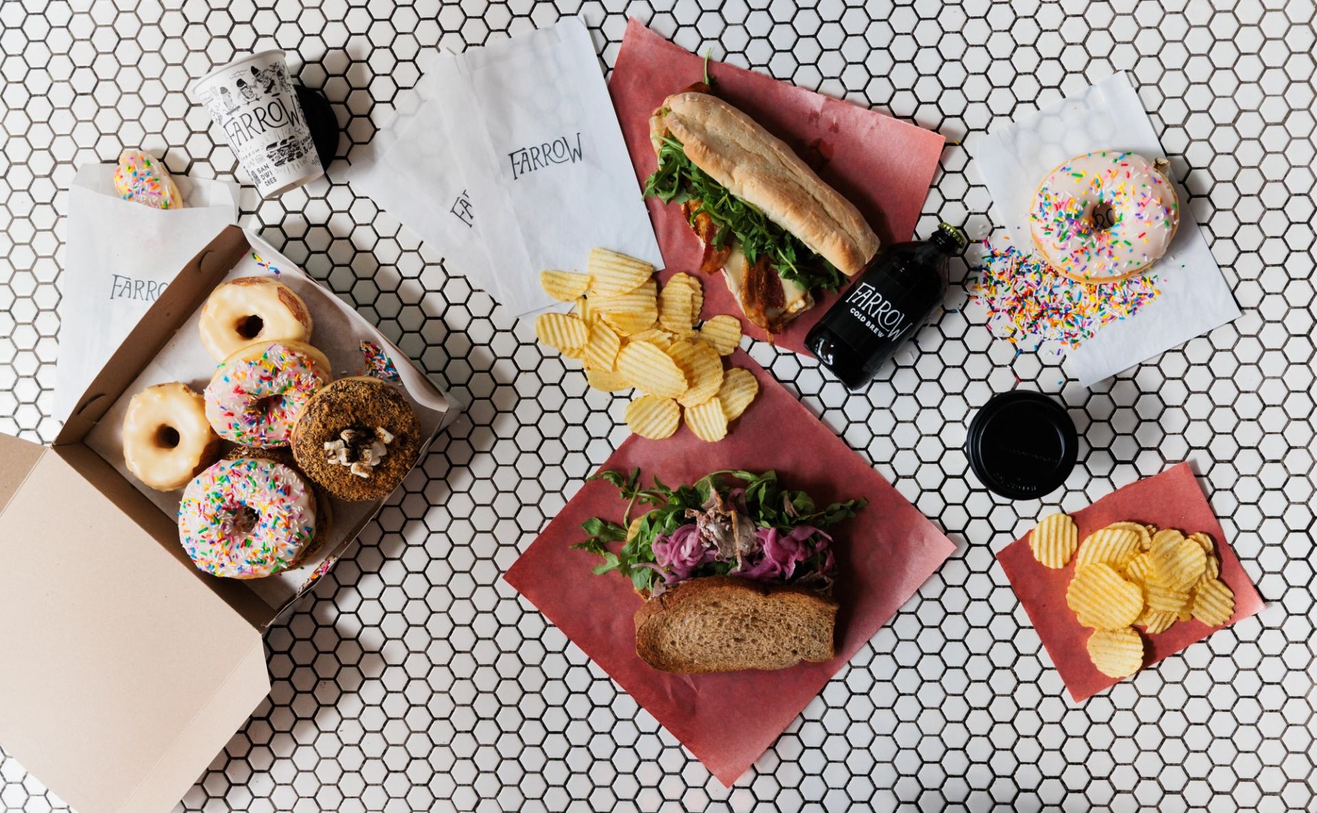 Sandwiches, doughnuts, potato chips and drinks arranged on a hexagonal tile counter.