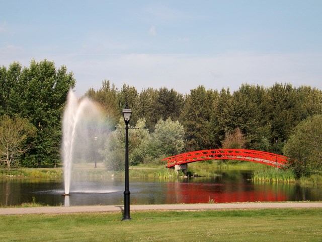 Park pond with red bridge, fountain, and lamppost surrounded by trees and blue sky.
