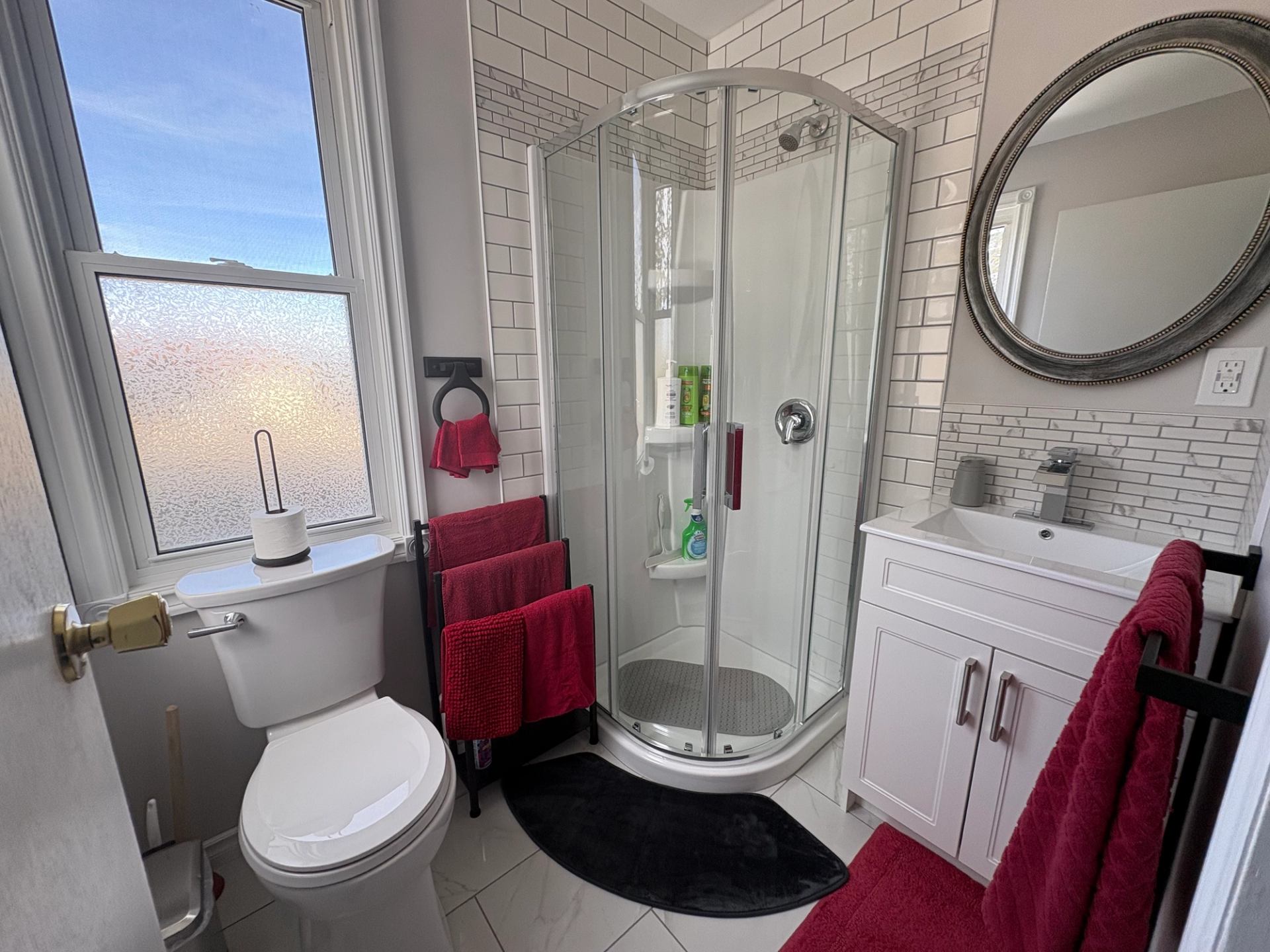 Small bathroom with corner shower, white tiles, and red towels on racks and vanity.