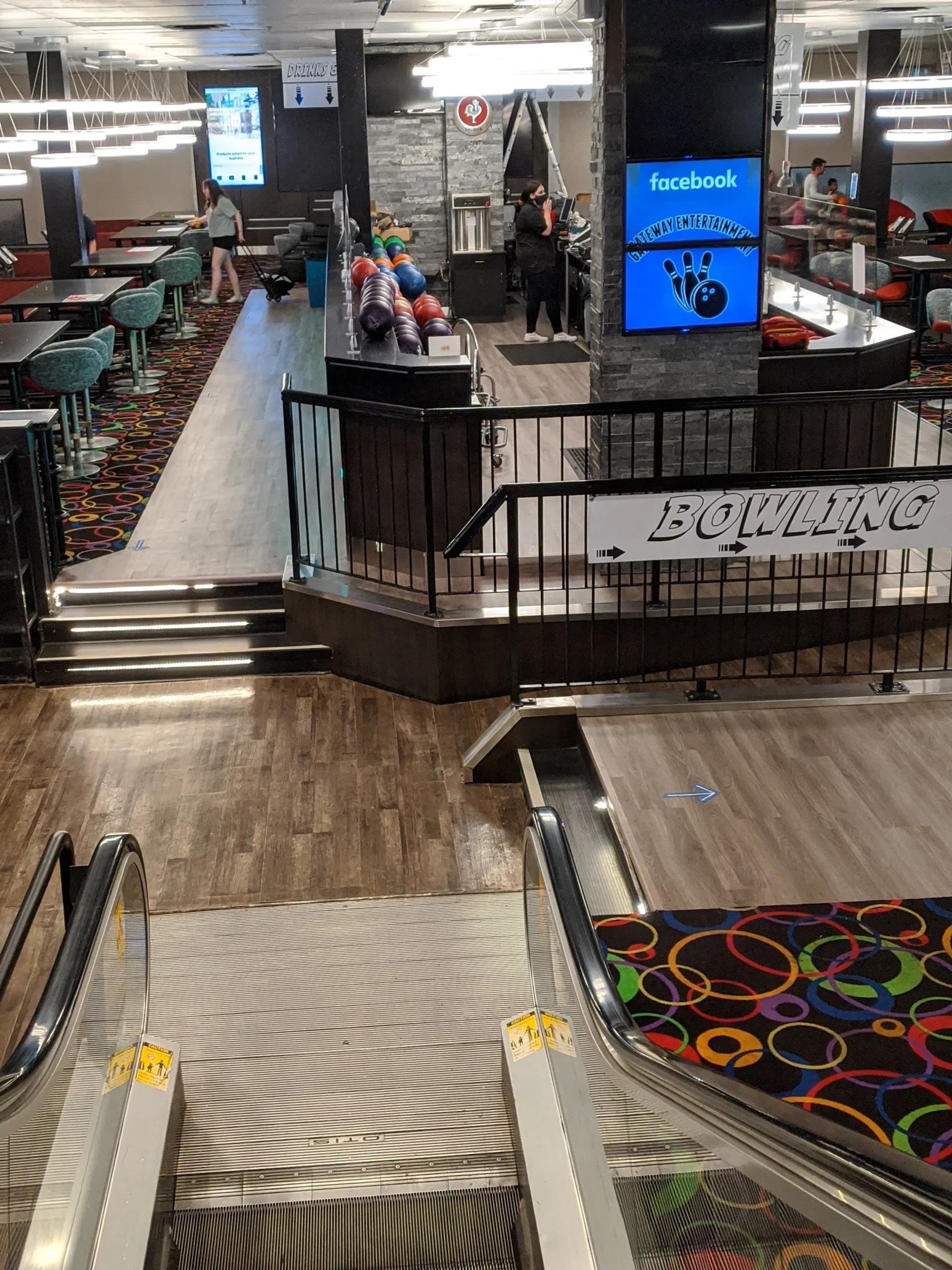 Bowling alley interior with escalator, colourful carpet, ball racks and seating area.
