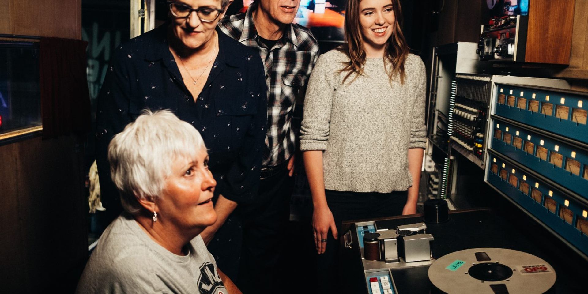 Group of people stand around recording equipment and tape reels in a control room.
