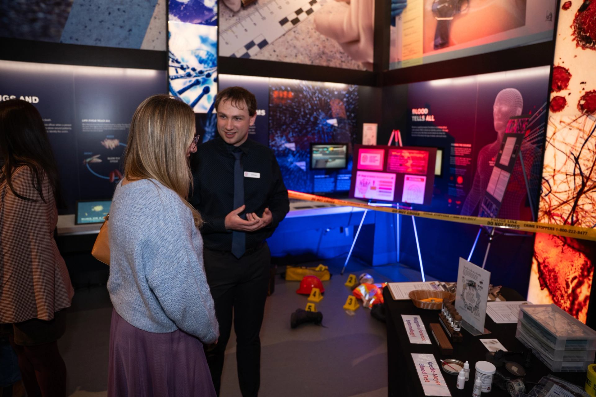 Museum guide speaks with a visitor beside a forensic crime scene exhibit.