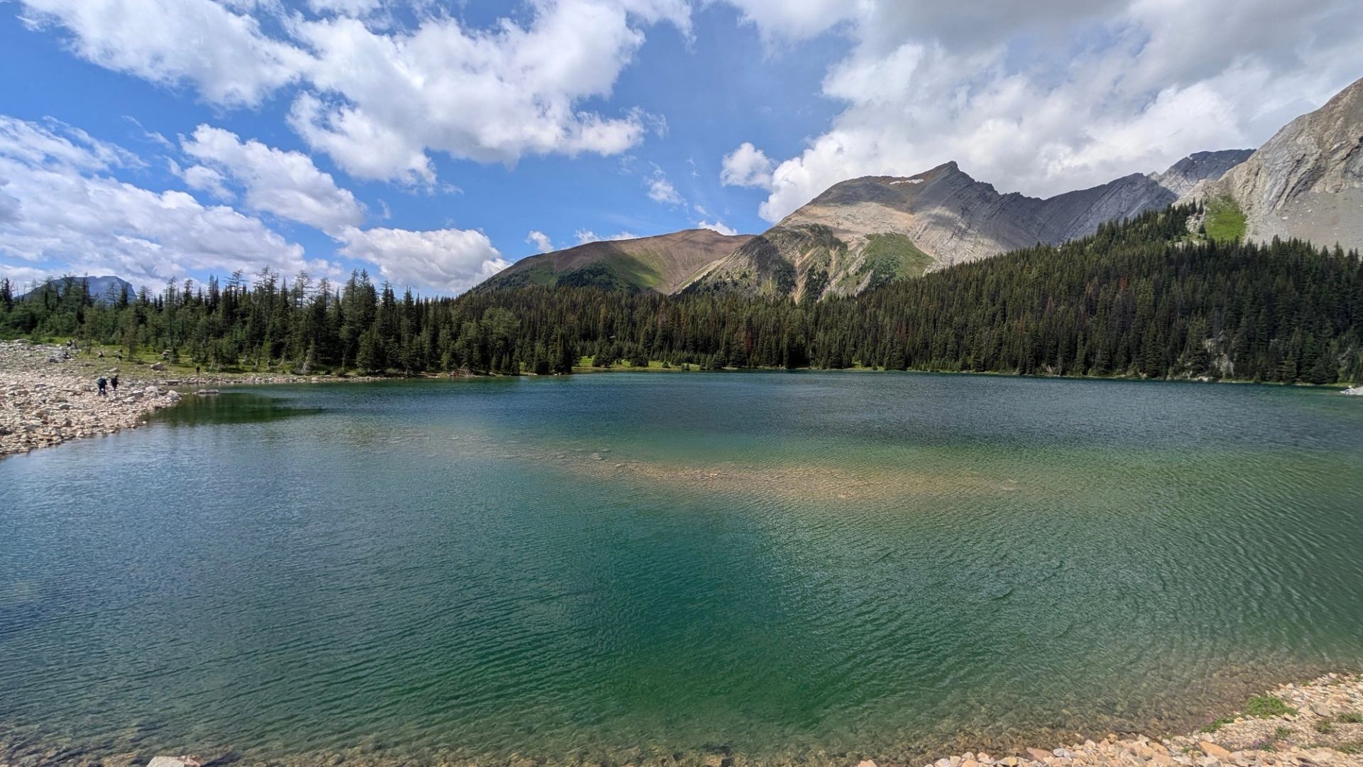Mountain lake with clear green water, forested shoreline and rocky peaks under clouds.