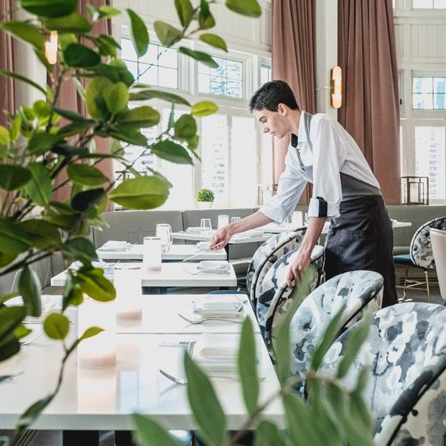Person arranging plates and napkins on a long table in a bright, elegant restaurant