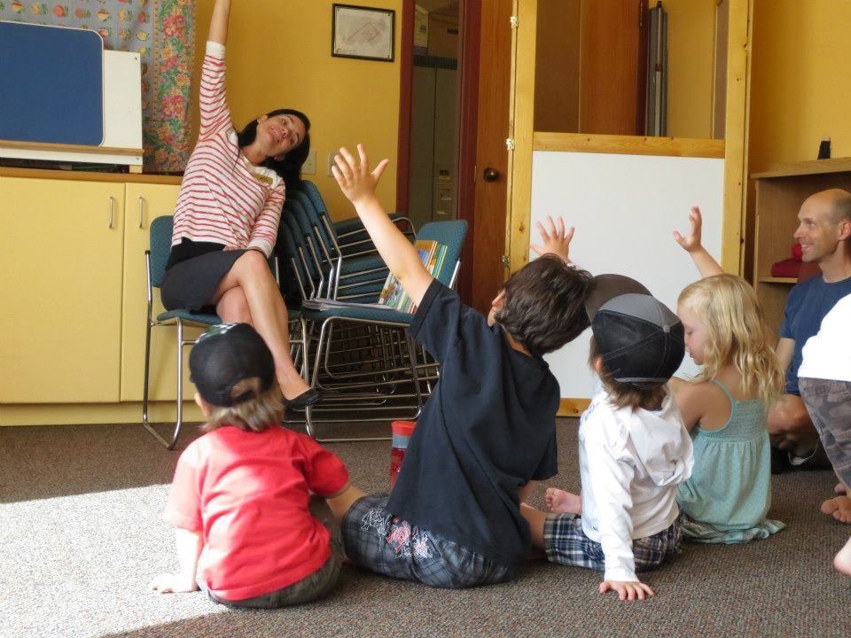 Children sitting on classroom floor while woman reads book and waves to them.
