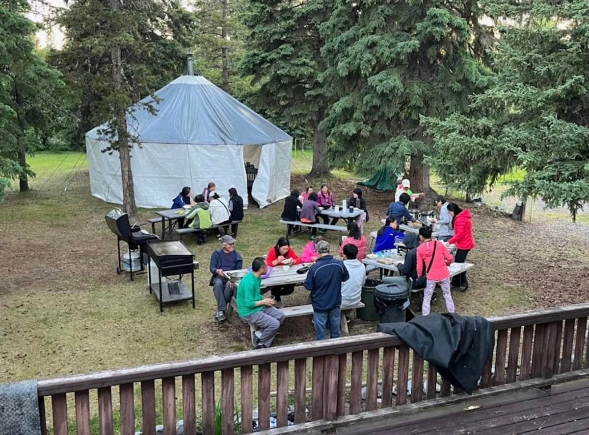 People gather around picnic tables beside a white tent among evergreen trees.