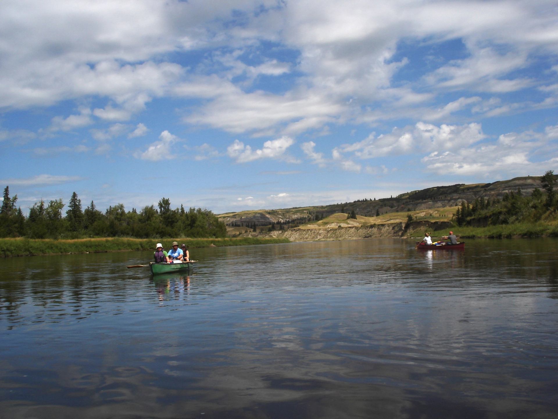 Canoes on calm river with forested banks and hills under blue sky.