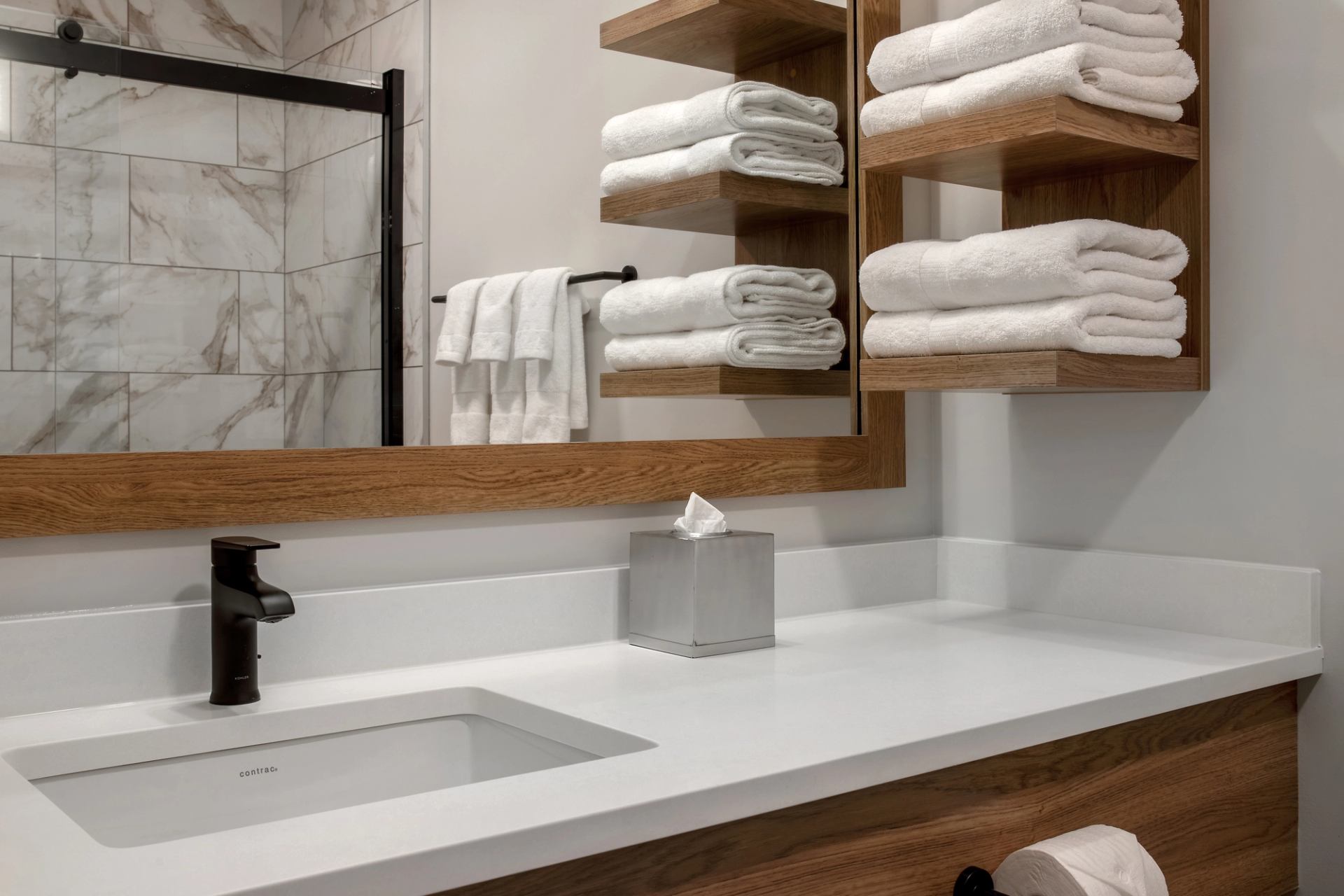 Bathroom vanity with white countertop, folded towels on wooden shelves, and glass shower.