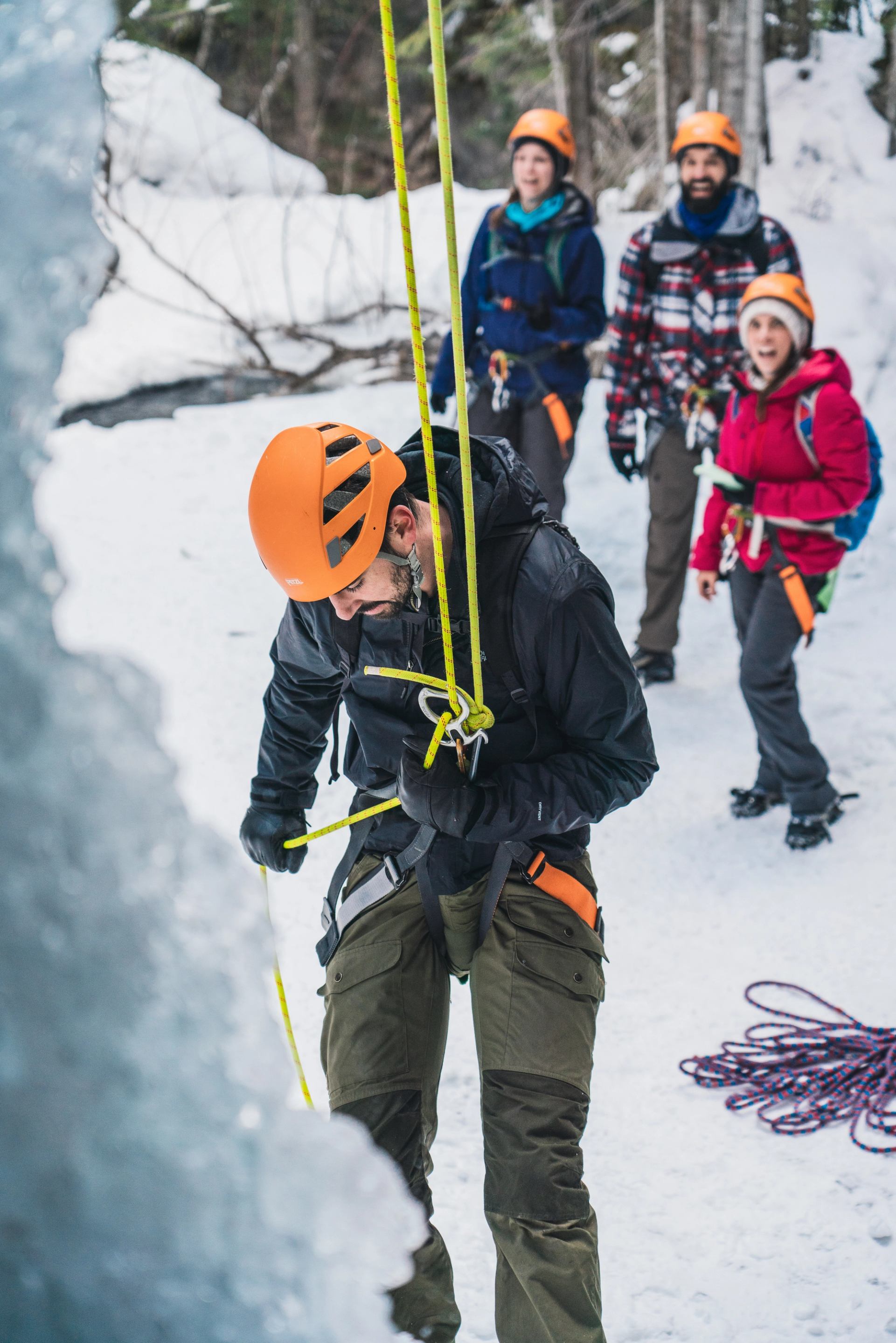 Group practicing rope setup on snow near icy canyon wall with climbing gear.