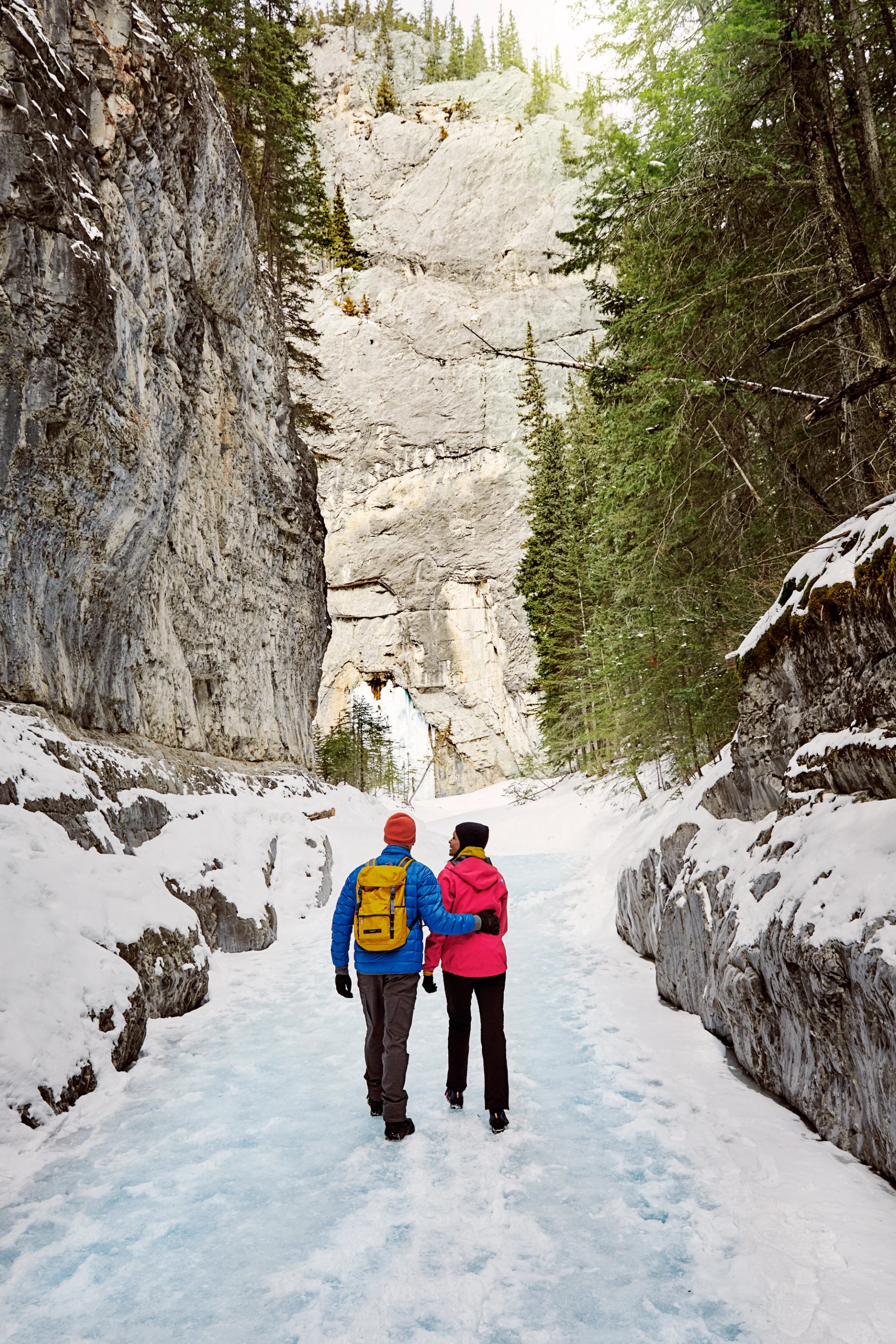 A couple walks arm-in-arm on a frozen river through a snowy canyon toward a rock arch and icefall.