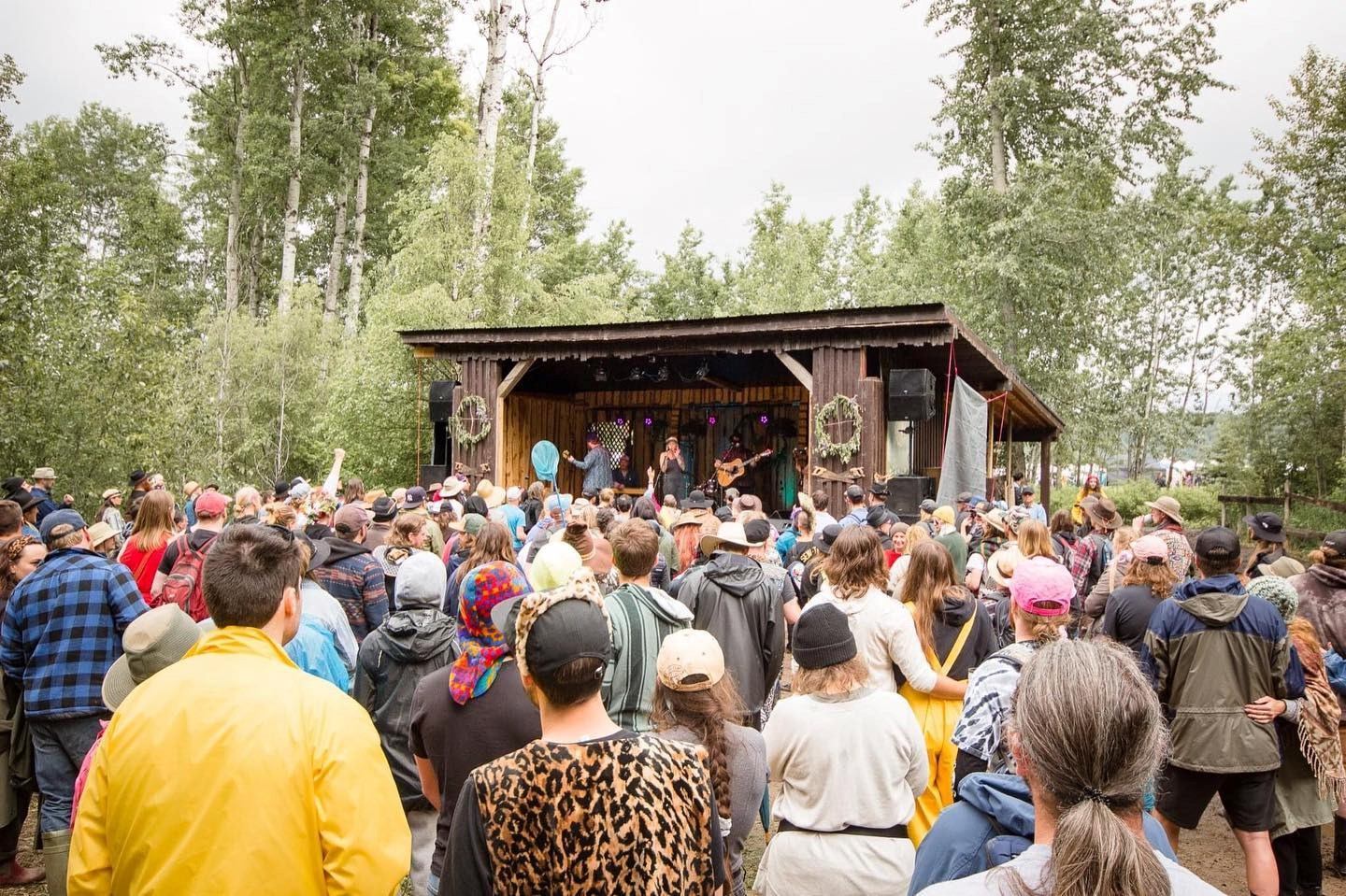 Crowd standing in a wooded clearing watching a band perform on a small outdoor stage at the fair.