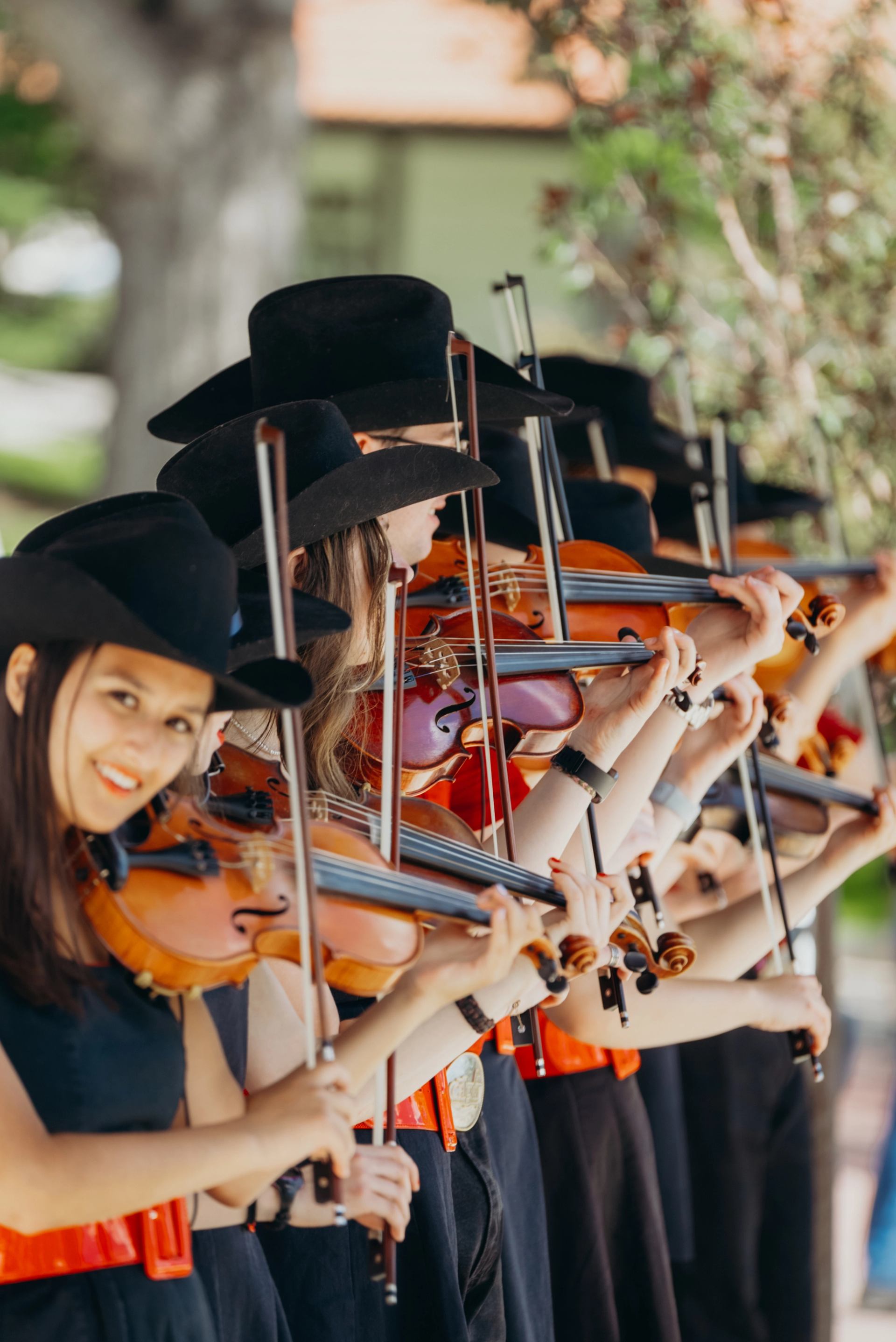 Musicians wearing cowboy hats and red belts play violins in a synchronized outdoor performance.